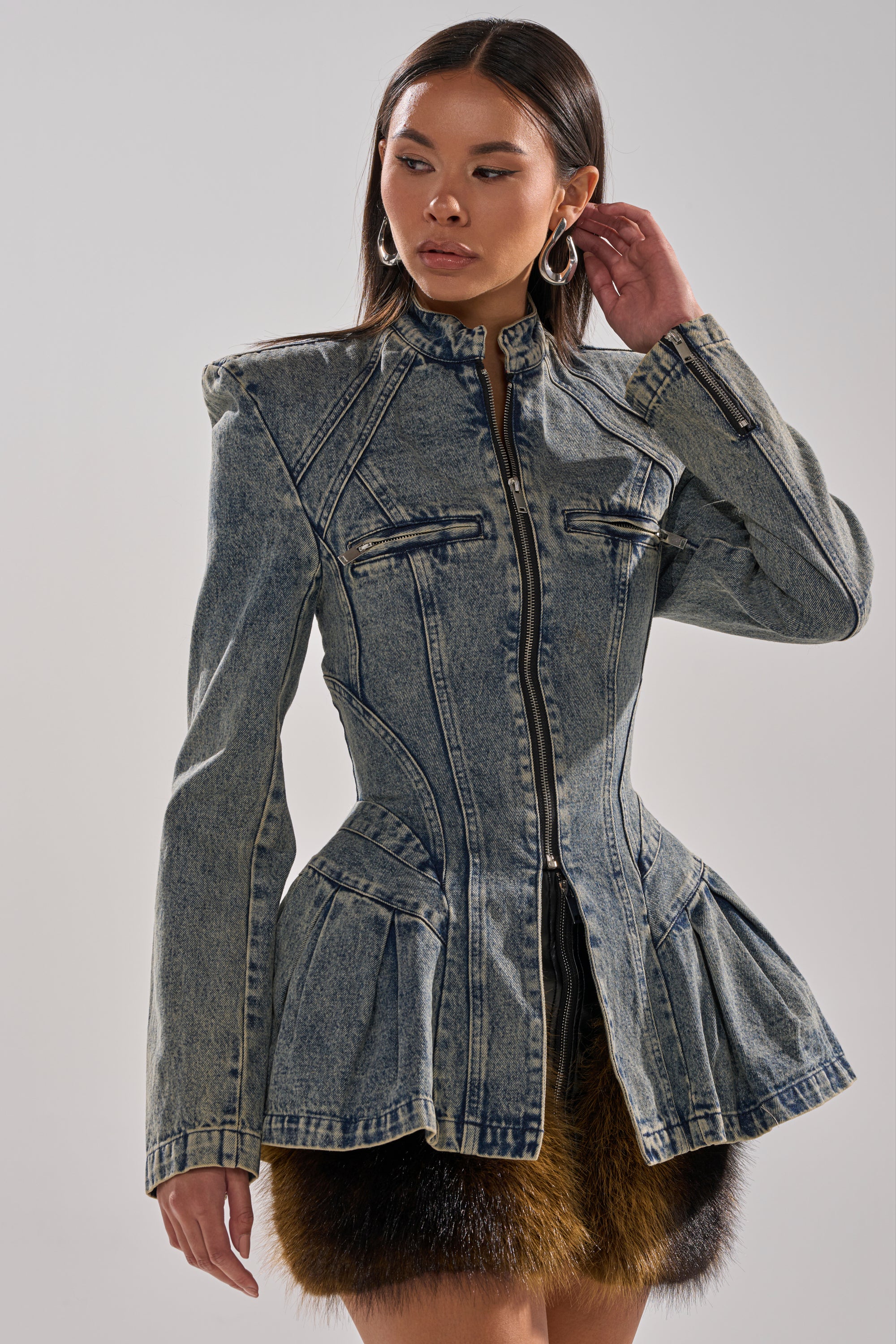 A woman models the MALLORCA PEPLUM DENIM JACKET, featuring a corset-style fit, high collar, zipper closure, prominent seams, and a peplum flared hem. She accessorizes with hoop earrings and wears her long hair straight.