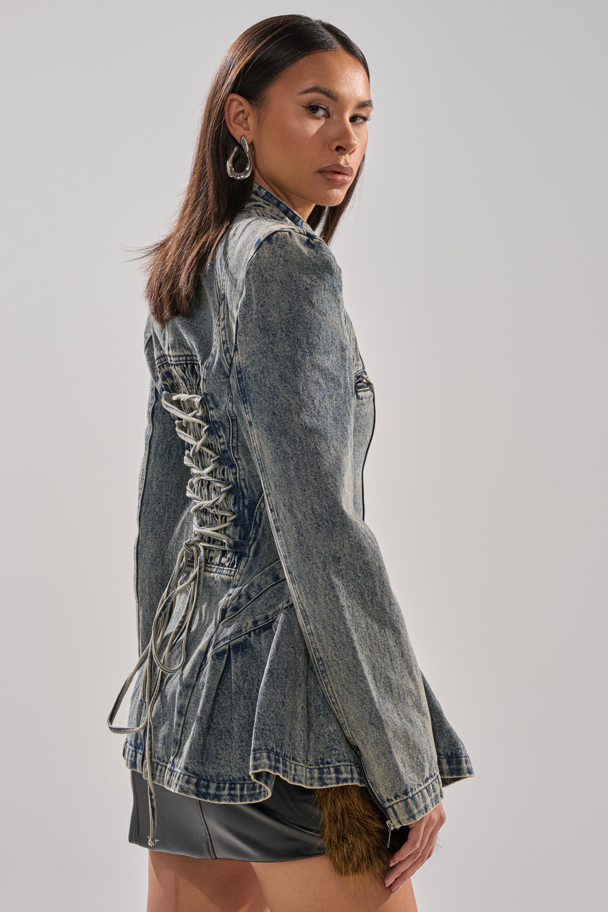 A woman with straight brown hair models the MALLORCA PEPLUM DENIM JACKET over a black leather mini skirt, looking over her shoulder against a plain light background.