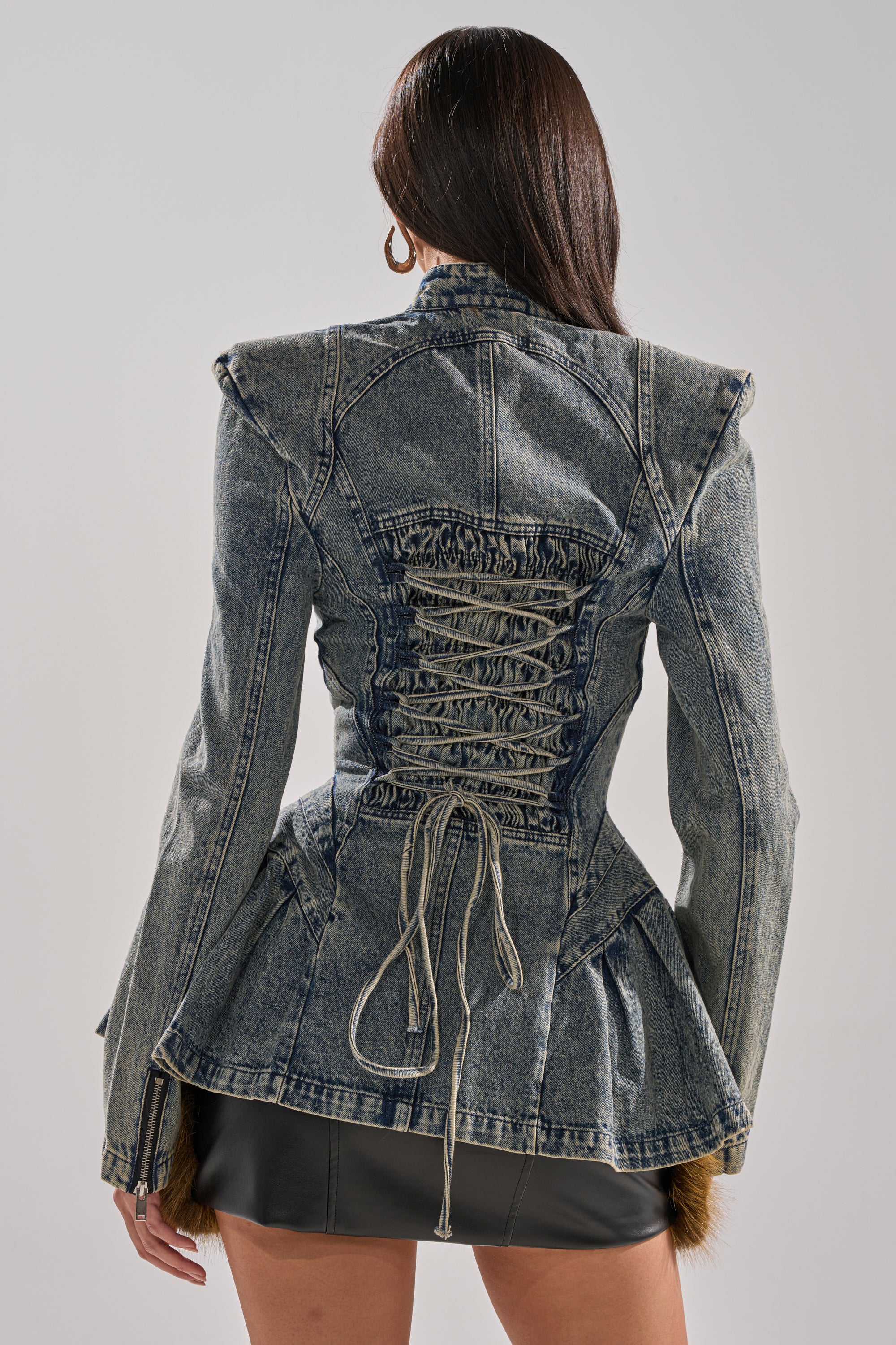 A woman with long, straight brown hair wears the MALLORCA PEPLUM DENIM JACKET, featuring corset-style lacing and a fitted silhouette, paired with a black mini skirt. She is seen from behind against a plain background.