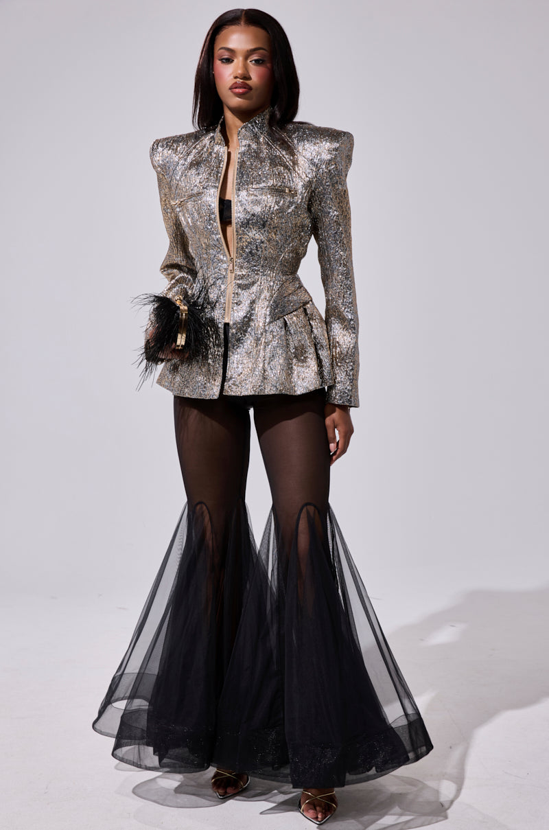 A woman stands confidently in the BRONZE BROCADE BLAZER with feather details and black sheer flared pants, paired with pointed heels. She has straight hair, bold makeup, and poses against a plain light background.
