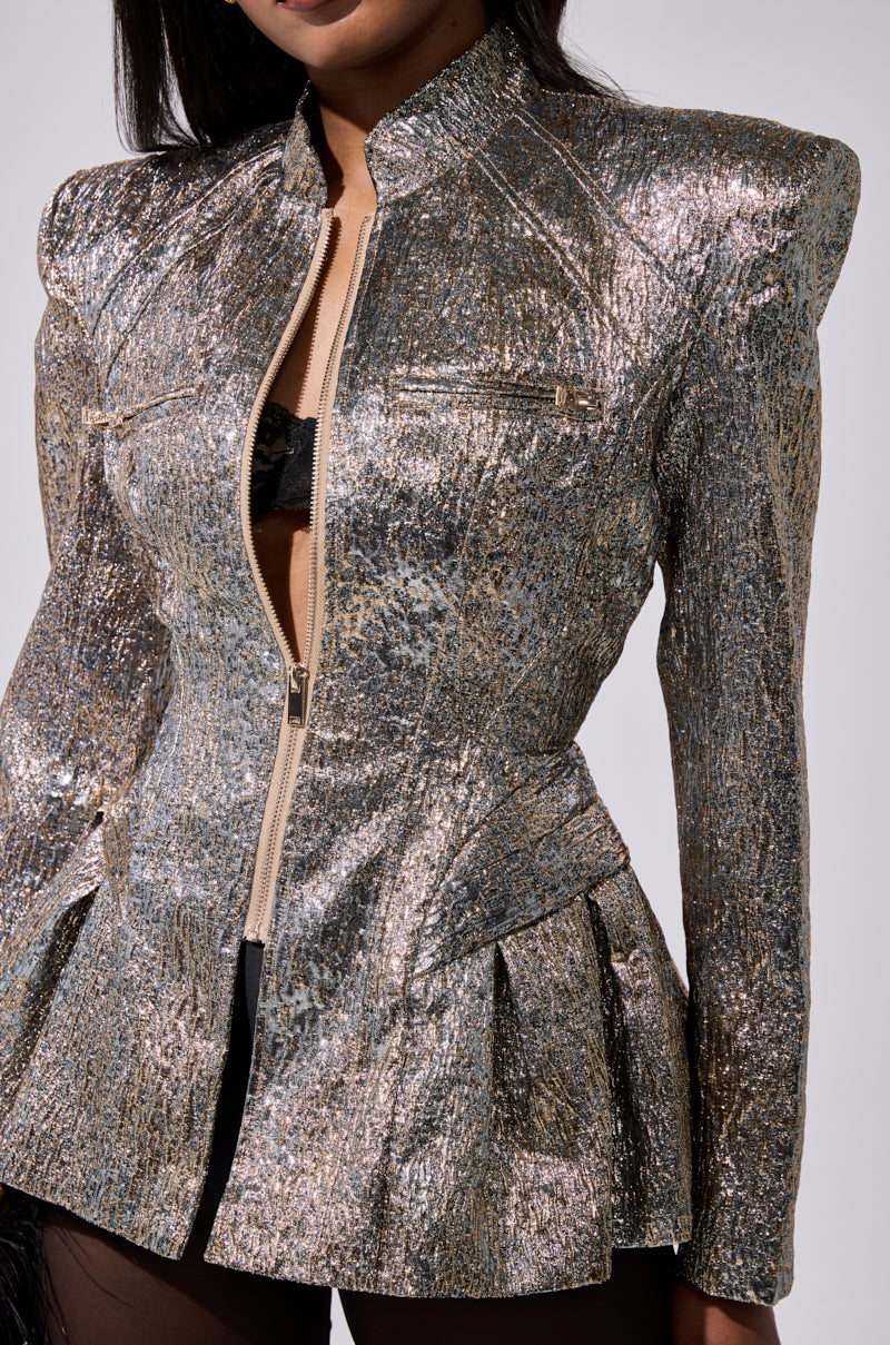 A woman wears the BRONZE BROCADE BLAZER, featuring strong shoulders, a zip-up front, and subtle pleats. Its textured, shimmering pattern is slightly open to reveal a black bra underneath.