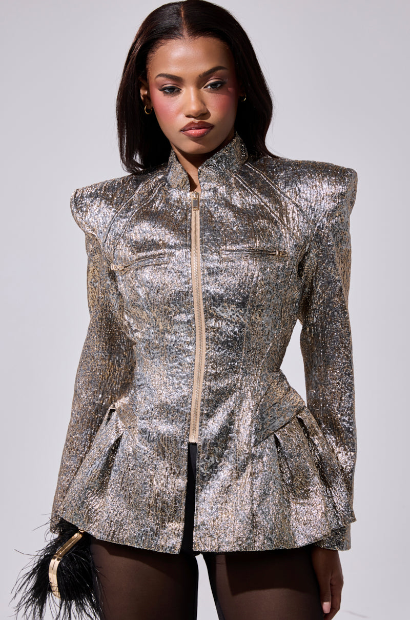 A woman stands facing forward, wearing the BRONZE BROCADE BLAZER with structured shoulders and a front zipper. She has straight dark hair, wears dark tights, and holds a black feathered accessory.