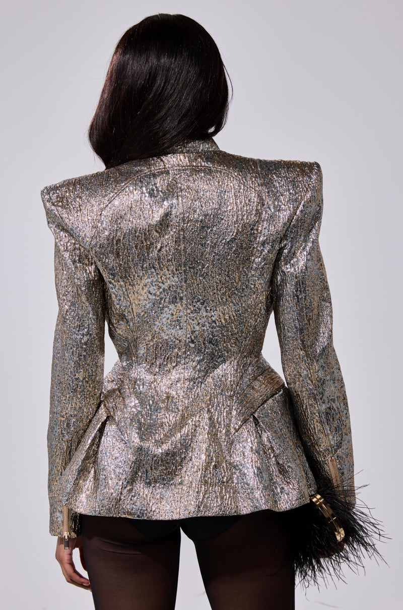 A person with long dark hair wears the BRONZE BROCADE BLAZER, featuring silver and gold metallic textures and structured shoulders, paired with black sheer tights. Facing away, they hold a black feathery accessory in their left hand.