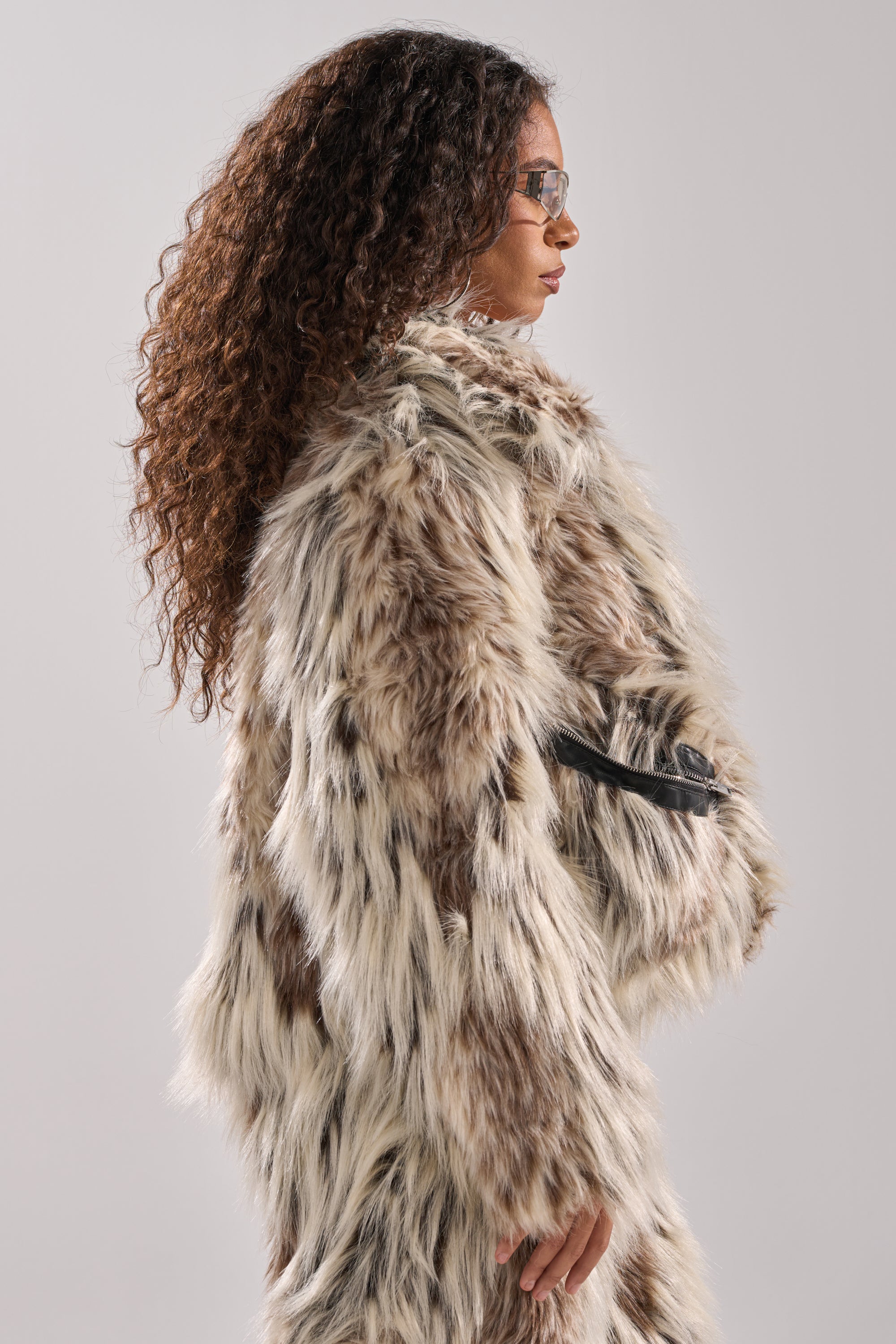A woman with long, curly brown hair and sunglasses models the PARTICULAR TASTE FAUX FUR coat with an oversized collar, standing in profile against a light gray background.