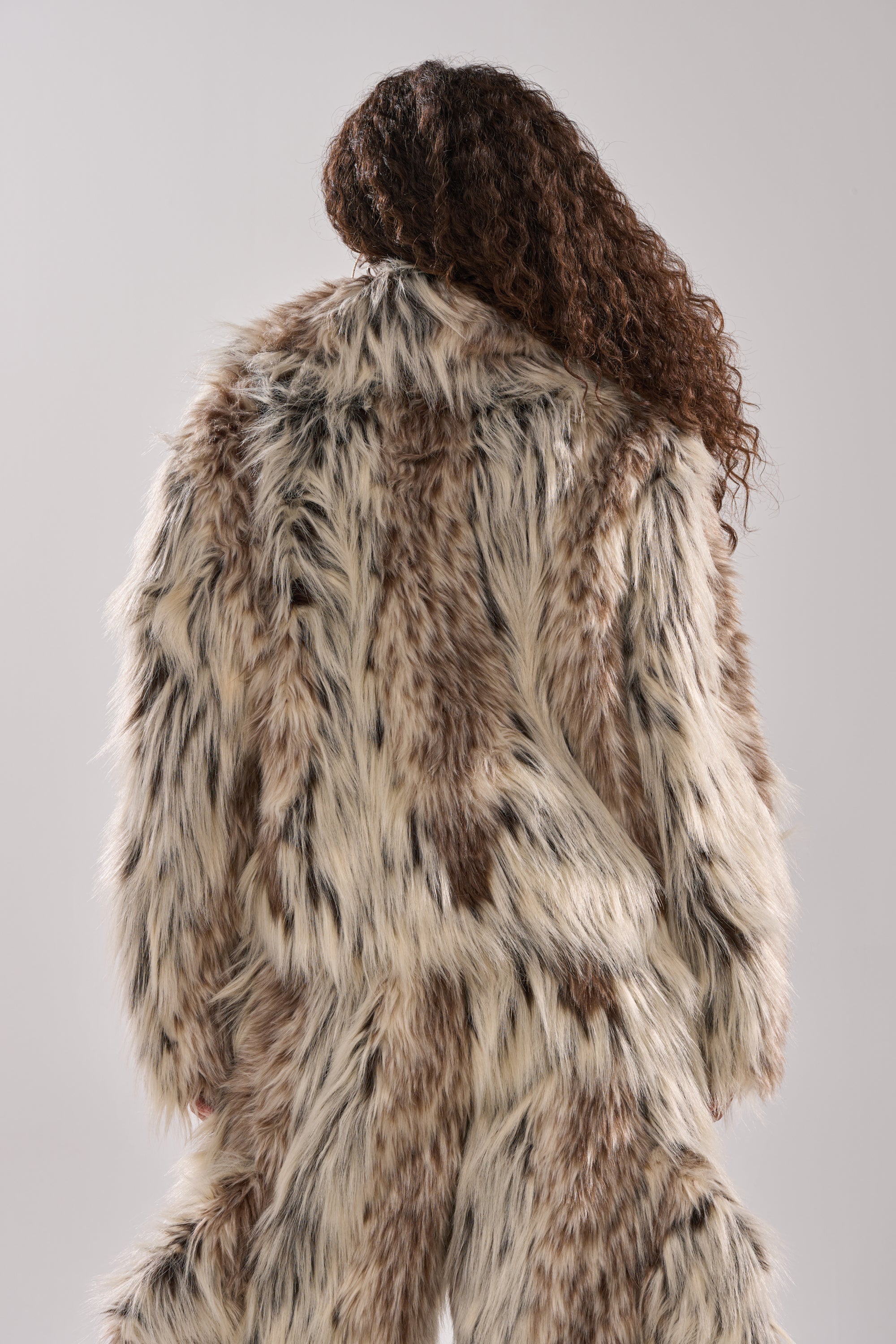 Person with long, curly hair wears the PARTICULAR TASTE FAUX FUR coat and matching pants, featuring an oversized collar and a blend of cream, brown, and black hues. Shown from the back against a plain light grey background.