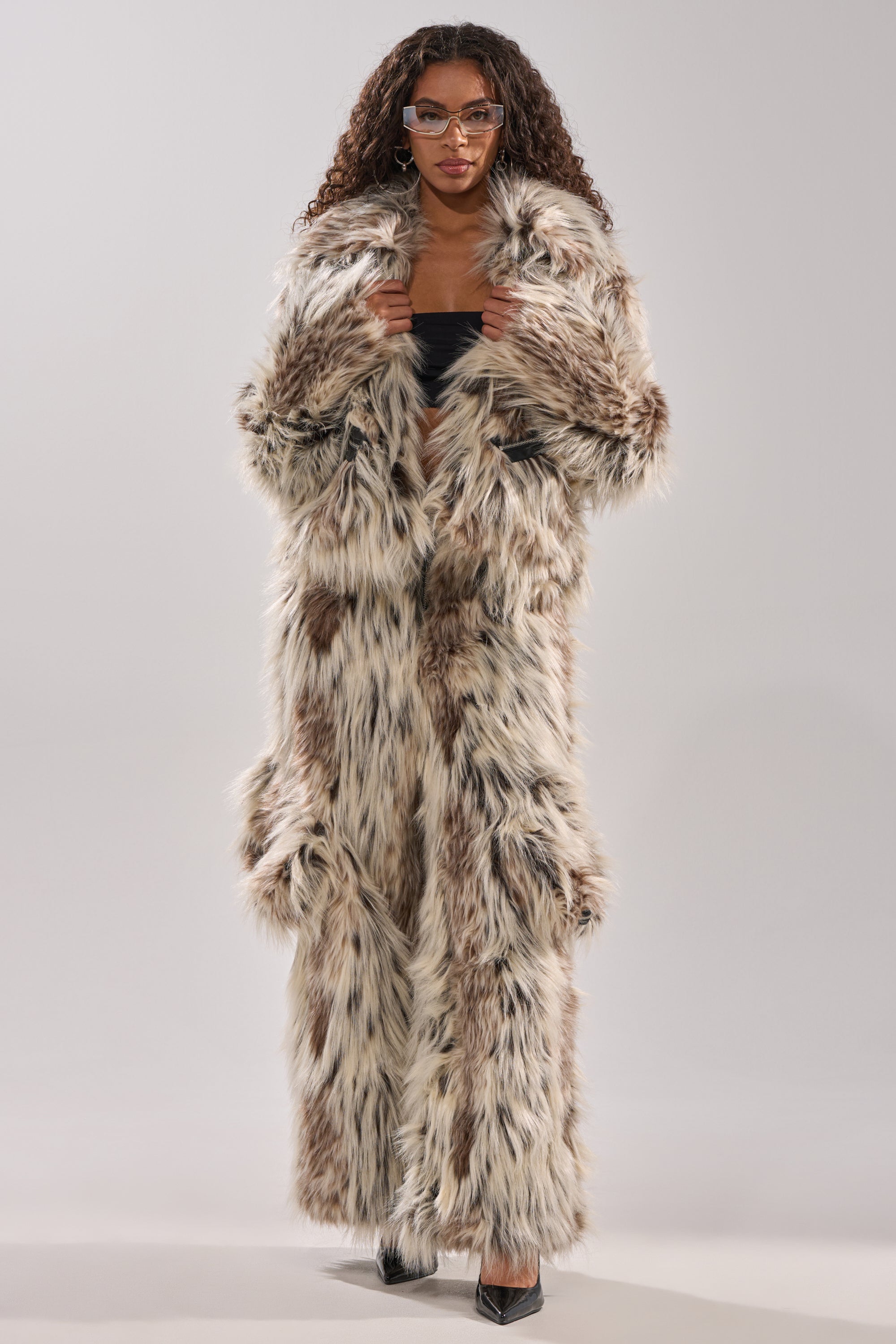 A woman wears the PARTICULAR TASTE FAUX FUR coat with an oversized collar in brown, white, and black hues, styled with a black top, black pants, heels, and clear-framed glasses against a plain light background.