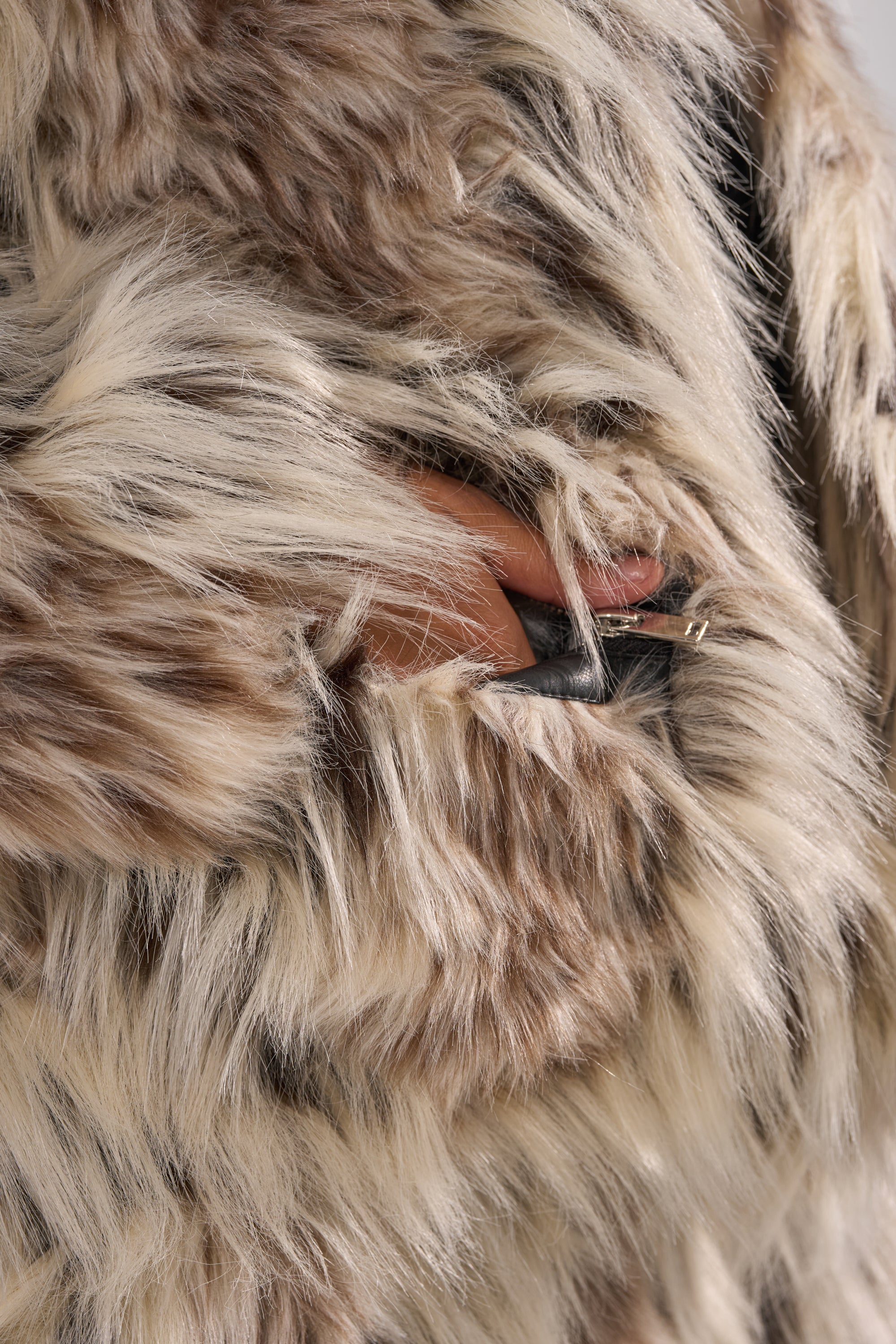 A hand holds the black zipper on the PARTICULAR TASTE FAUX FUR coat, featuring plush brown-and-white material and an oversized collar.