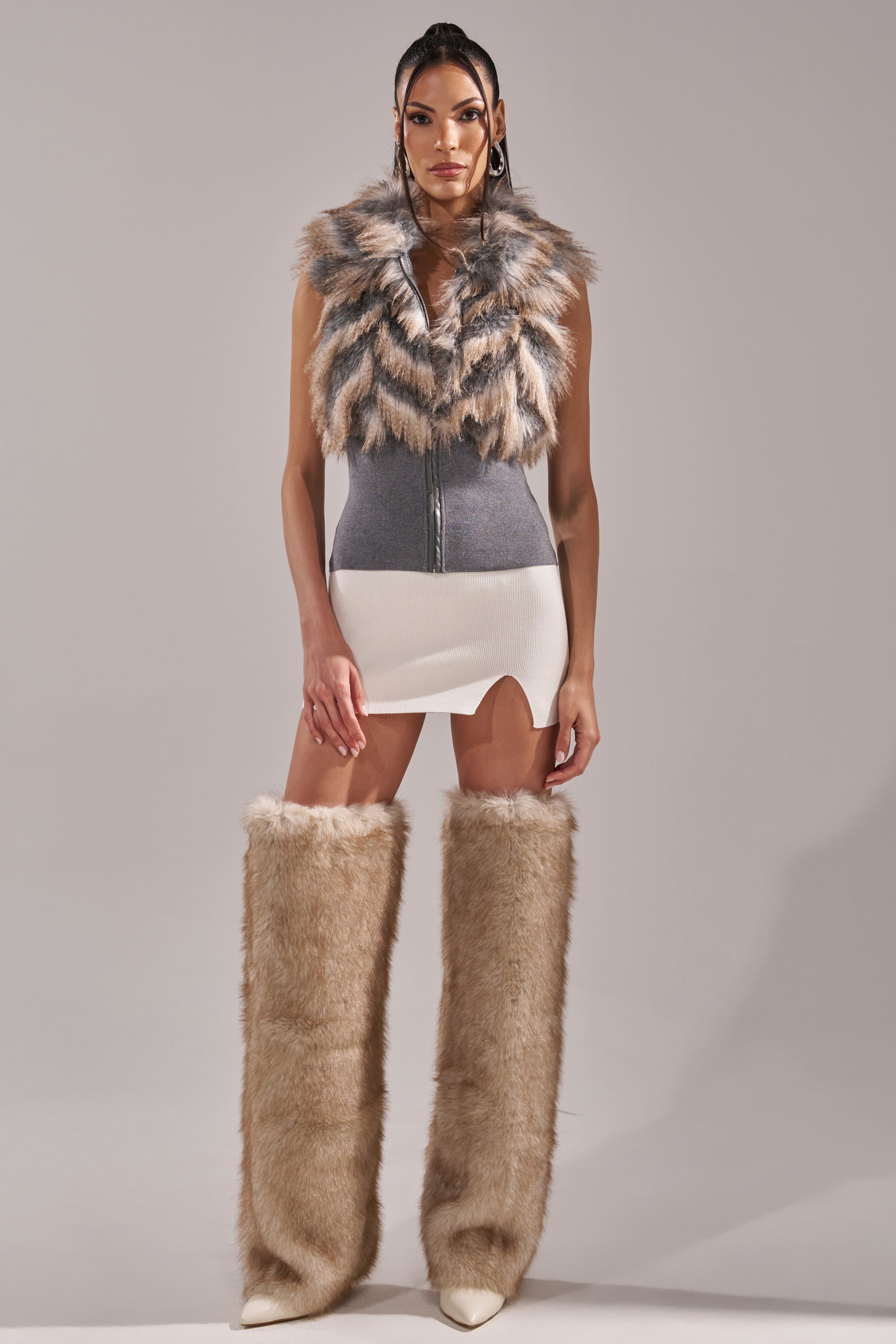 A woman with long, straight hair and a neutral expression faces forward wearing the NEW SENSATION FUR TOP—a gray and white sleeveless mini dress with a faux fur collar—paired with tan over-the-knee faux fur boots. 8