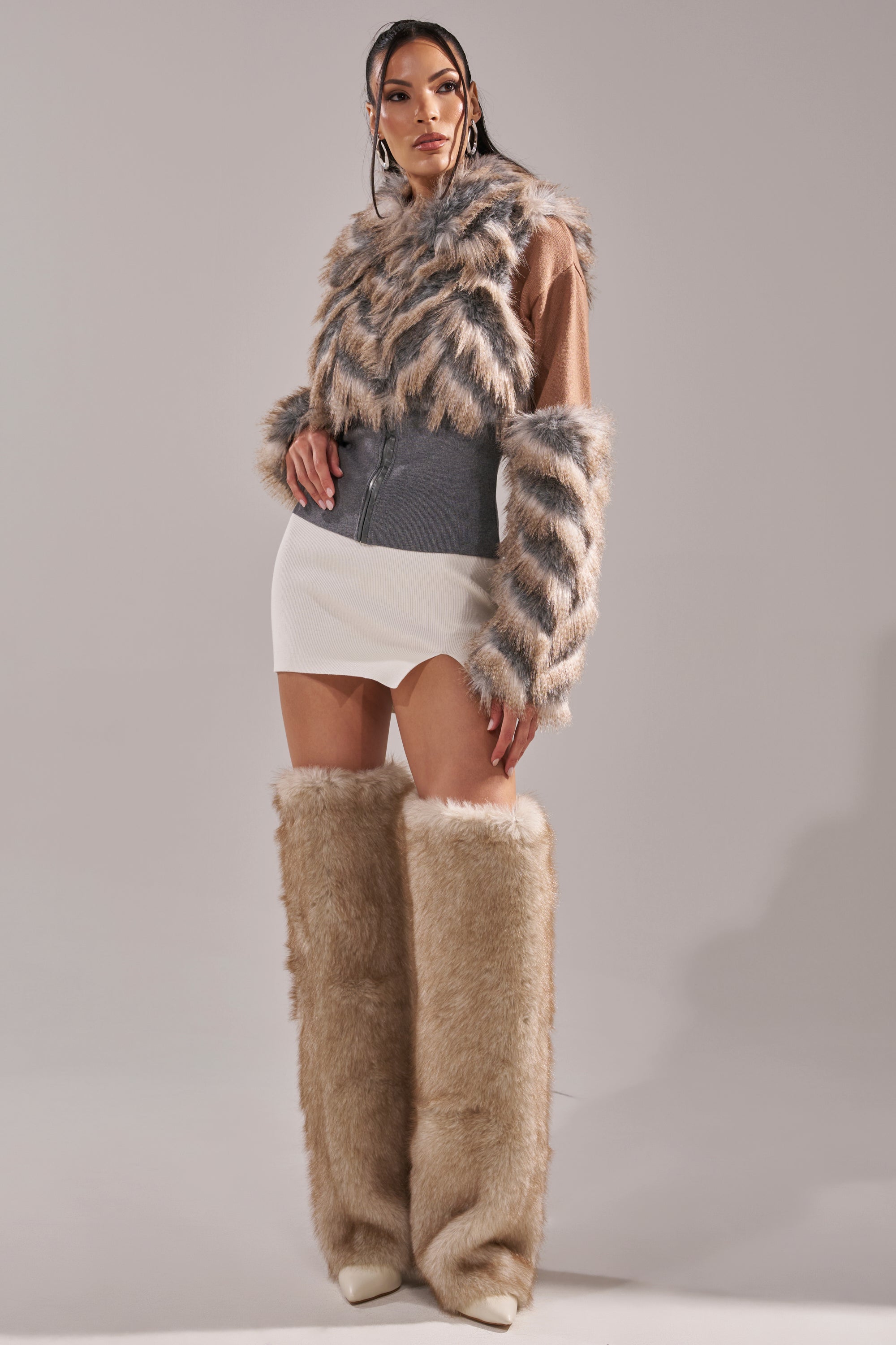 A woman poses in the AKIRA Label NEW SENSATION FUR TOP, paired with a white mini skirt and tall fluffy beige fur boots, against a plain light gray background. 10