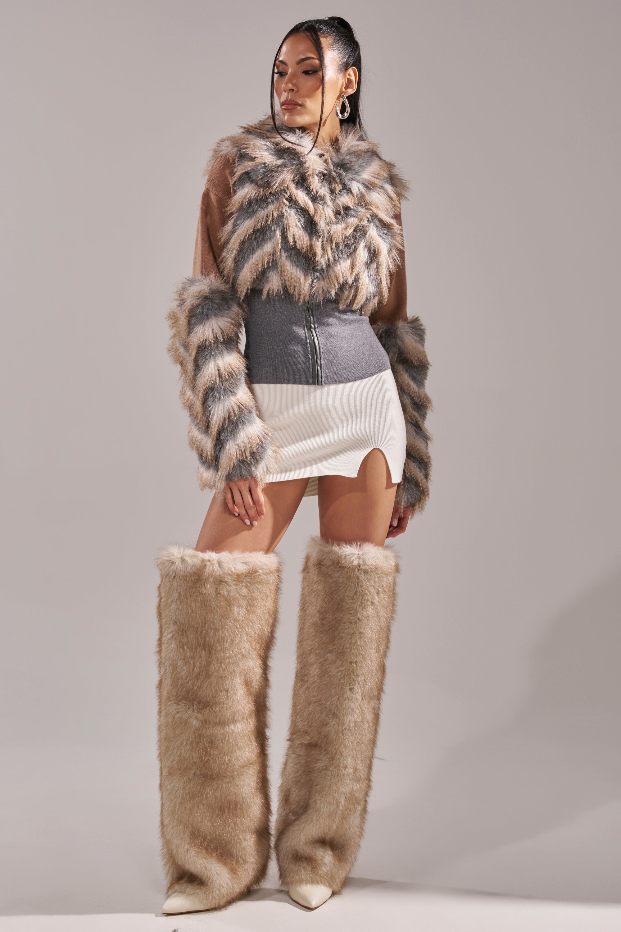 A woman models the NEW SENSATION FUR TOP—faux fur with an oversized collar and striped sleeves—paired with a gray corset, white mini skirt with front slit, and tall tan faux fur boots against a plain light background. 12