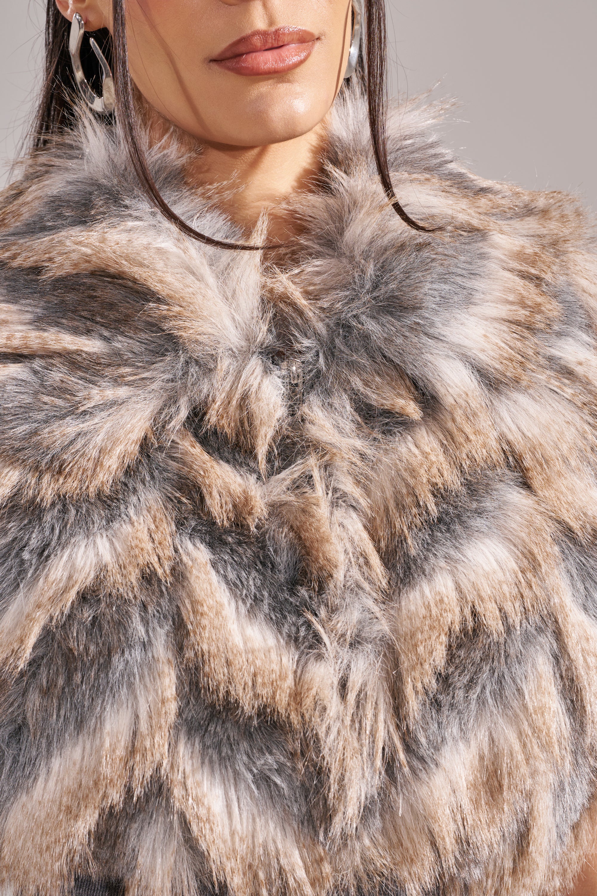 A woman models the NEW SENSATION FUR TOP, a chic tri-tone coat in gray, brown, and beige, with just her lips, chin, and earrings partially visible. 13