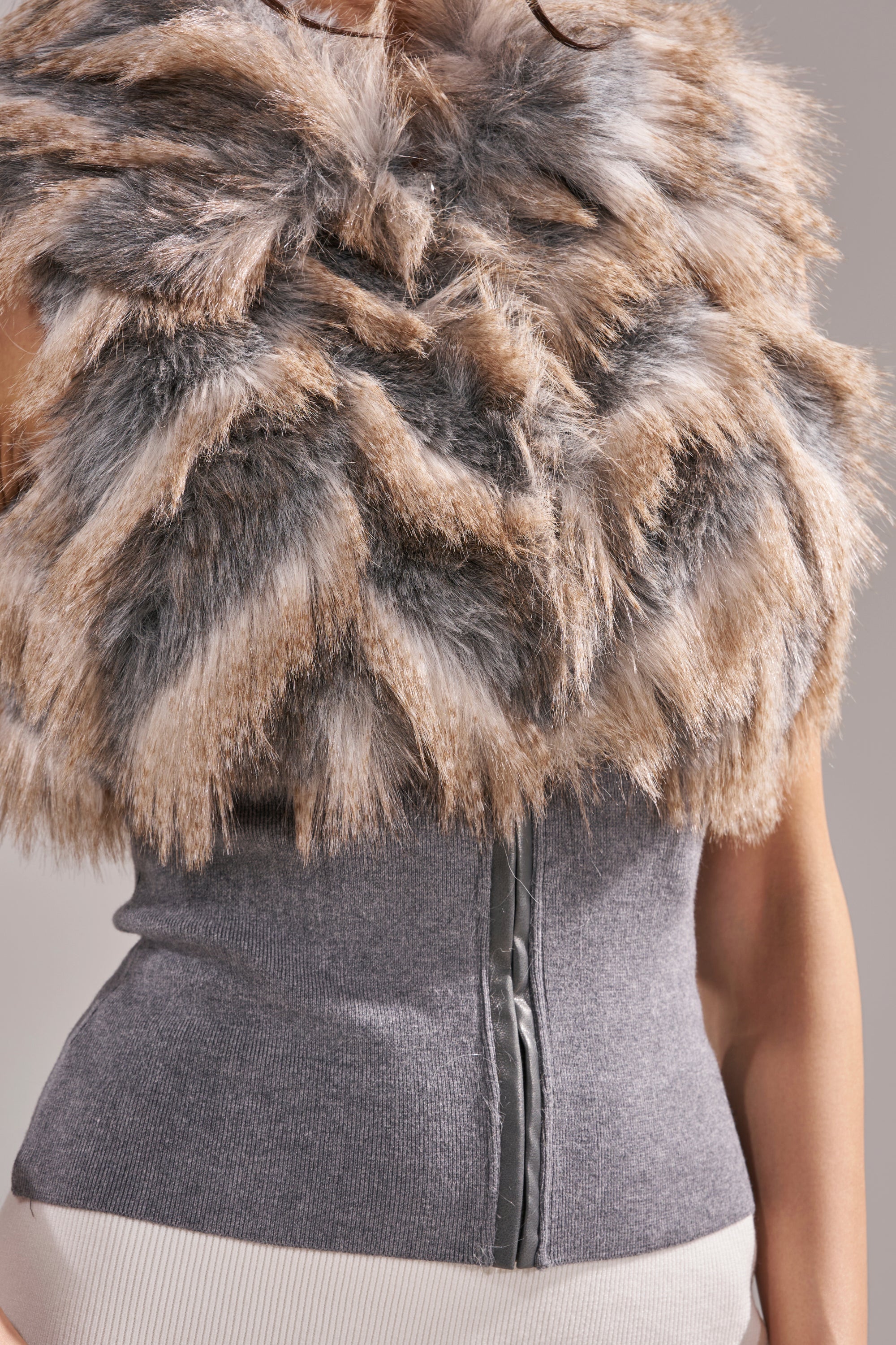 A person wears the NEW SENSATION FUR TOP, a tri-tone faux fur piece in brown, gray, and white, layered over a fitted sleeveless gray zip-up crop top. 14