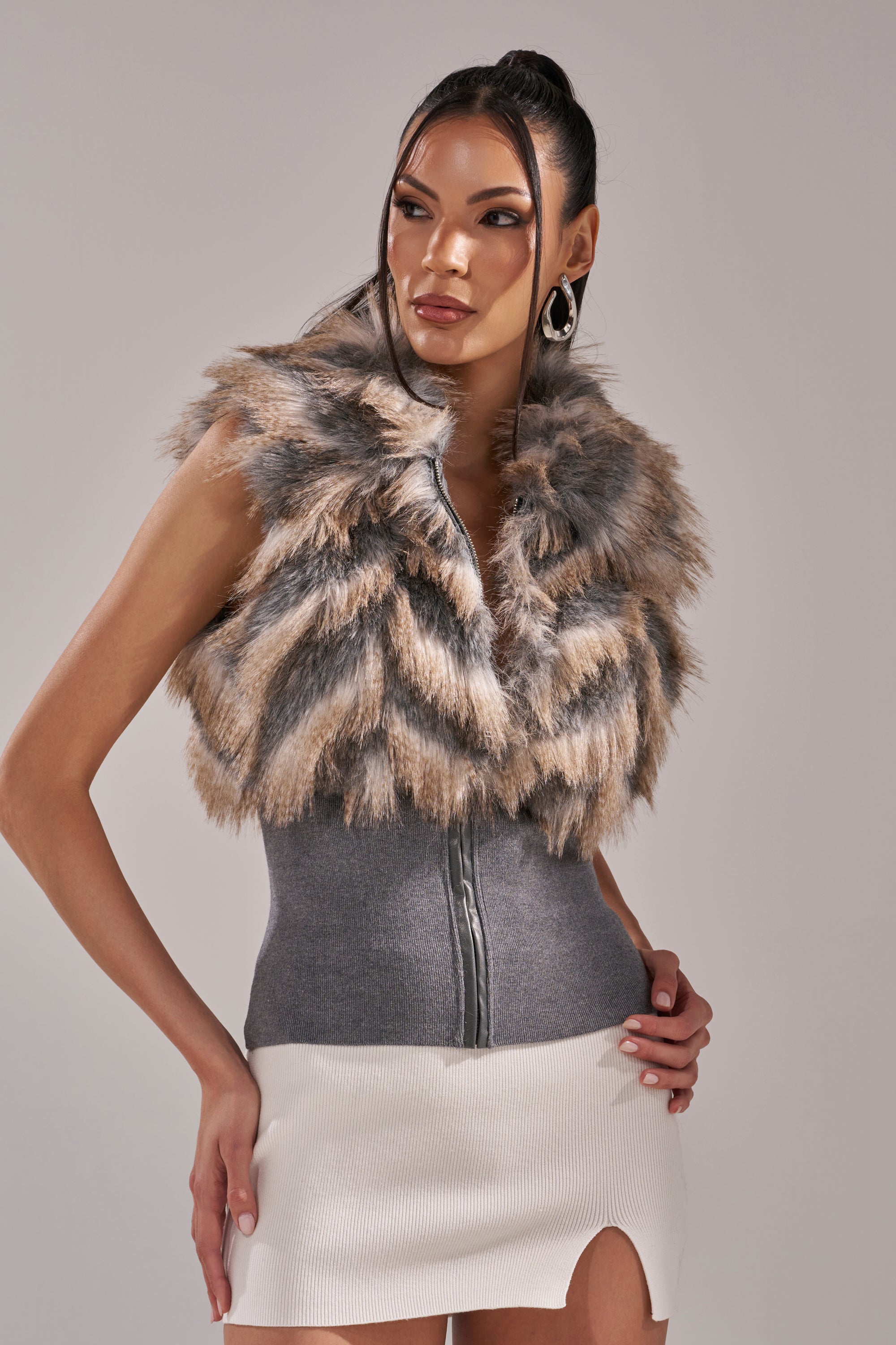 A woman wears the NEW SENSATION FUR TOP—an AKIRA Label sleeveless faux fur piece in gray, brown, and cream—layered over a fitted gray top and a white slit skirt, standing against a plain light background. 3