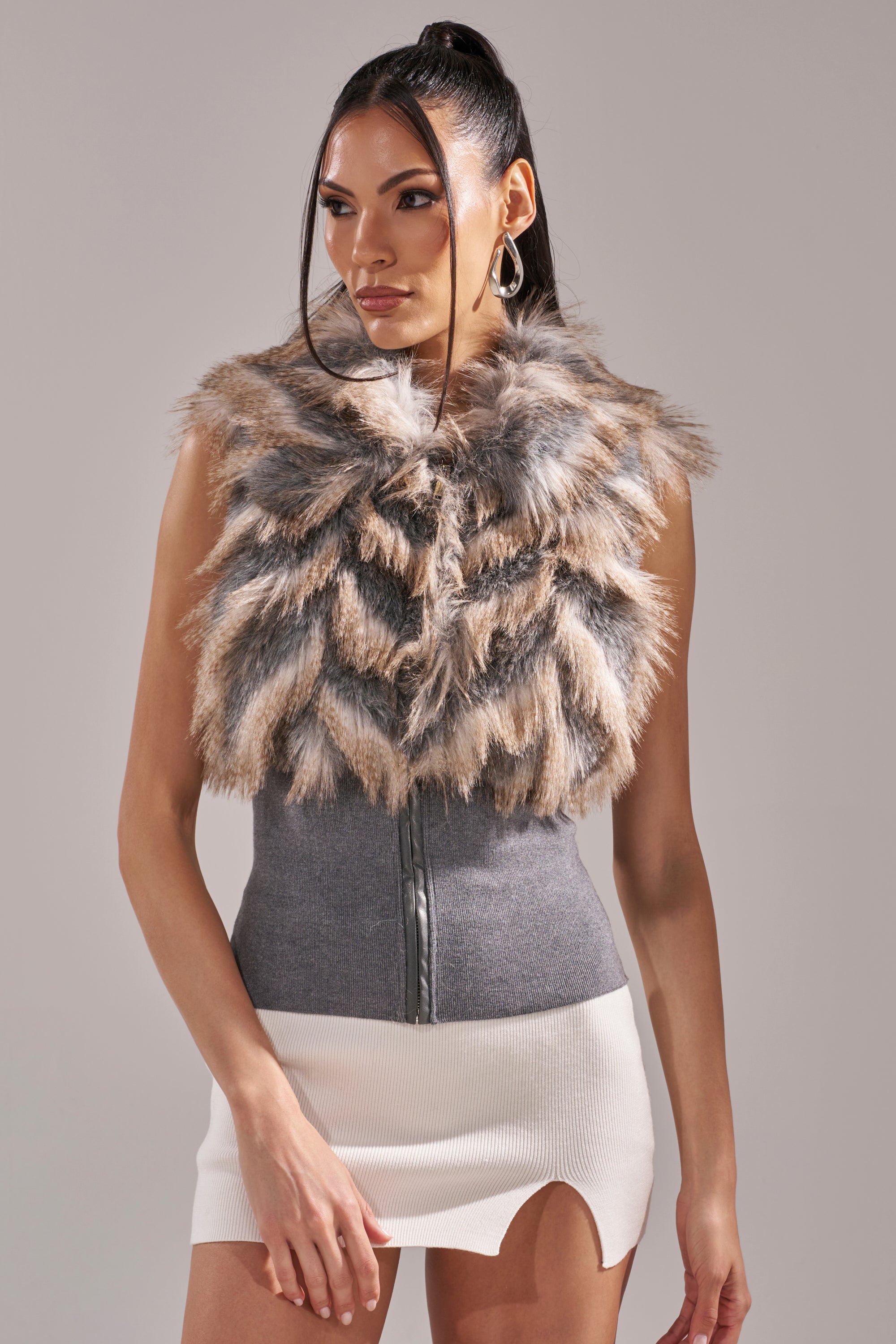 A woman with long dark hair in a high ponytail wears the NEW SENSATION FUR TOP, a sleeveless fitted gray faux fur piece with a central zipper, styled with a short white skirt featuring an asymmetrical hem against a plain background. 4