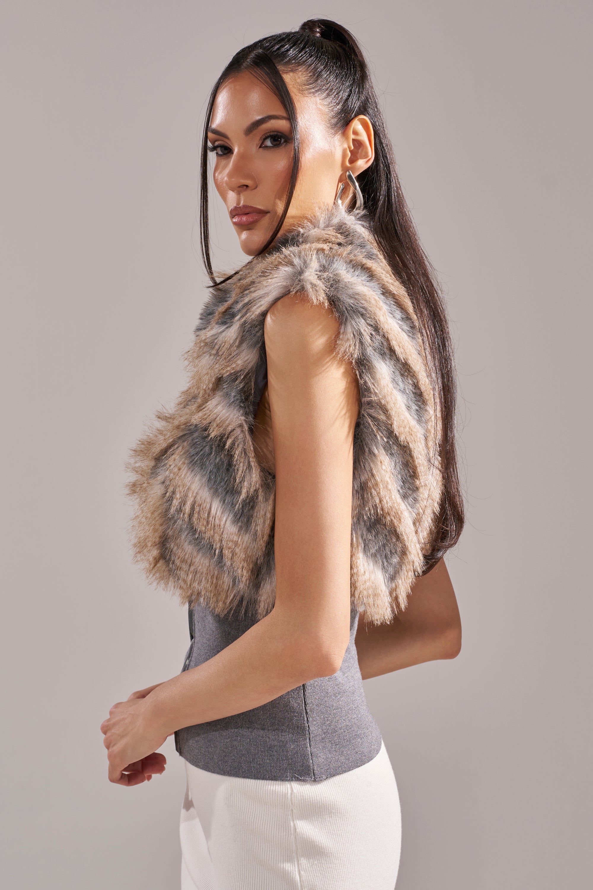 A woman with long, dark hair in a high ponytail models the NEW SENSATION FUR TOP—sleeveless with gray and beige stripes—layered over a gray top and white pants, standing against a plain light background. 6