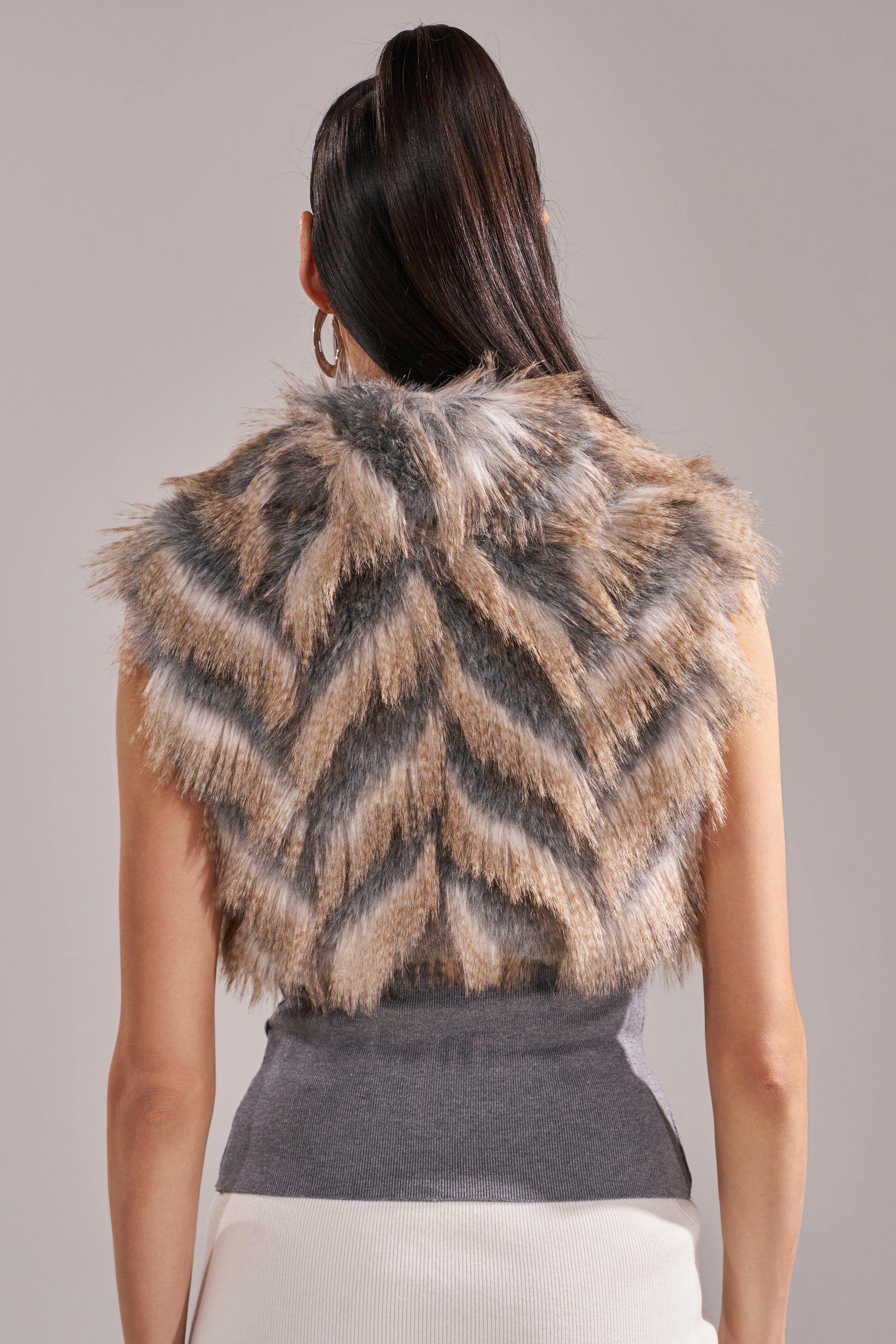 A woman with long dark hair in a high ponytail is seen from the back wearing the NEW SENSATION FUR TOP: a sleeveless chevron-patterned tri-tone (brown, black, and white) faux fur top layered over a grey shirt. 7