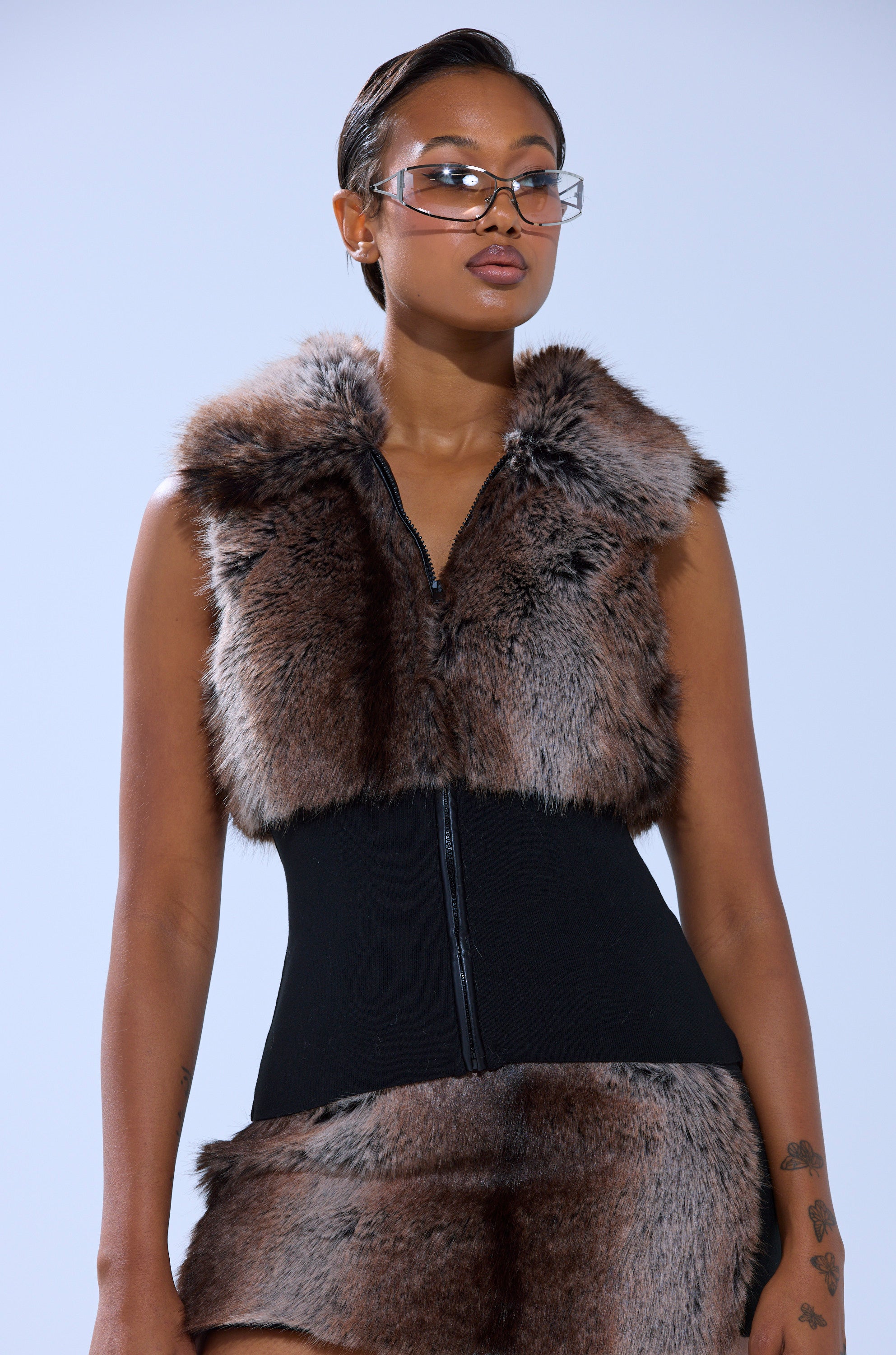 A person models the FEELS GOOD FUR BLOUSE—sleeveless with a ribbed elastic hem—over a black fitted outfit and faux fur skirt, completed with clear futuristic glasses, set against a plain light blue background.