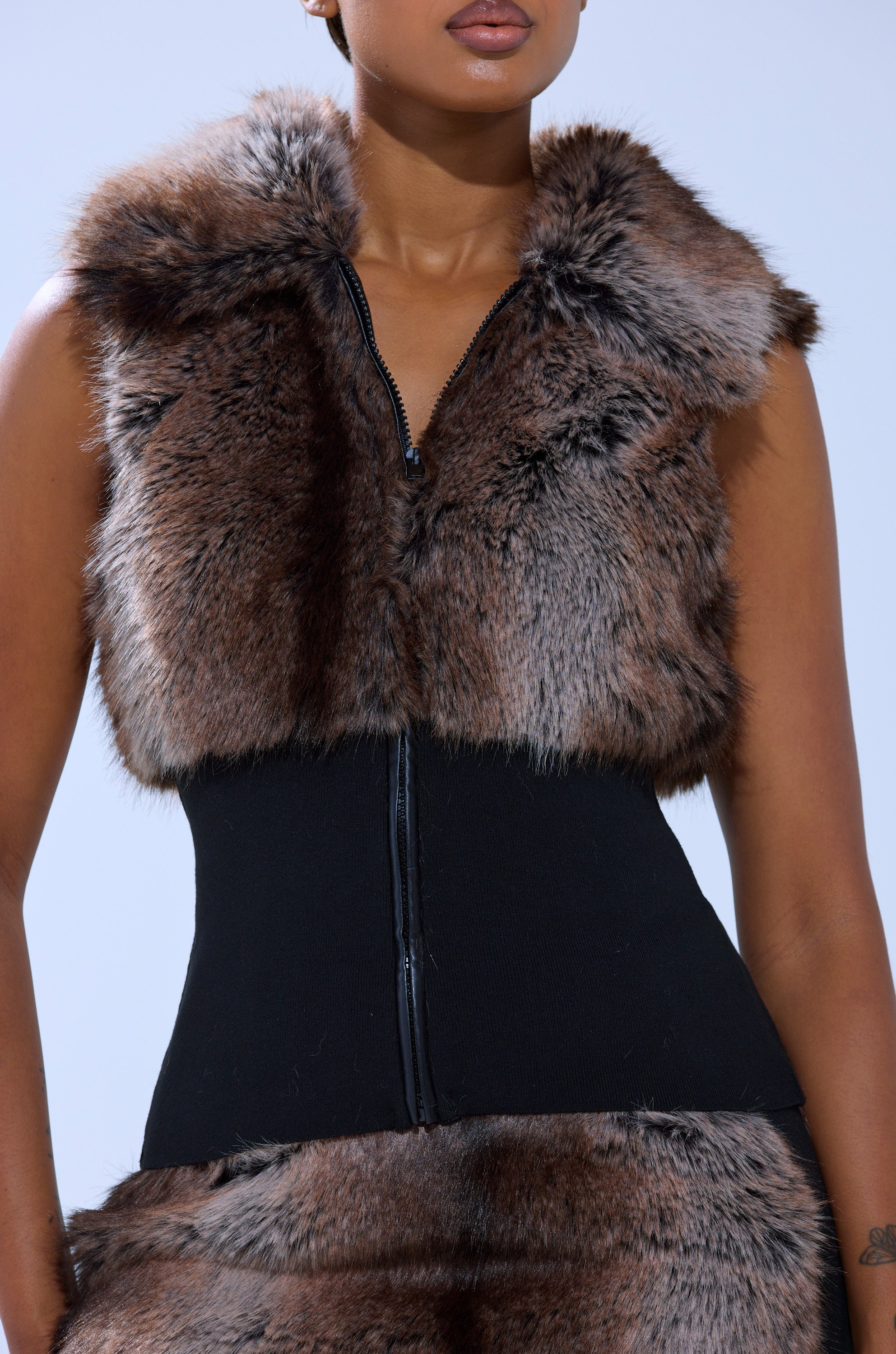 A person wears the FEELS GOOD FUR BLOUSE, a sleeveless fur top with a zipper, layered over a fitted black garment. The image is cropped to show just the torso and part of the arms.