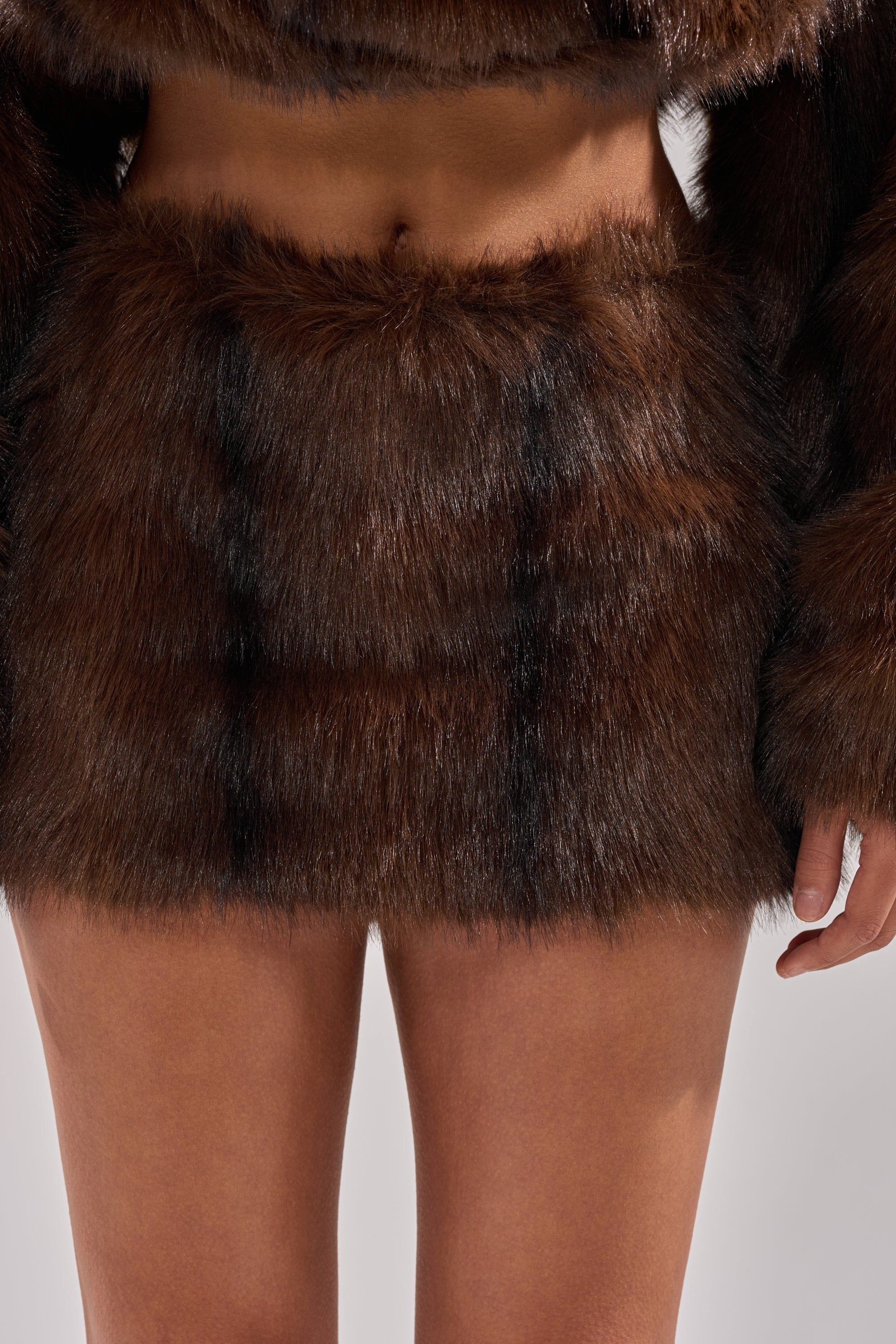 A close-up of a person wearing the COZY VIBES FUR MINI SKIRT in brown, paired with a matching faux fur top, showing their bare midriff and legs, with one arm partially visible. 1