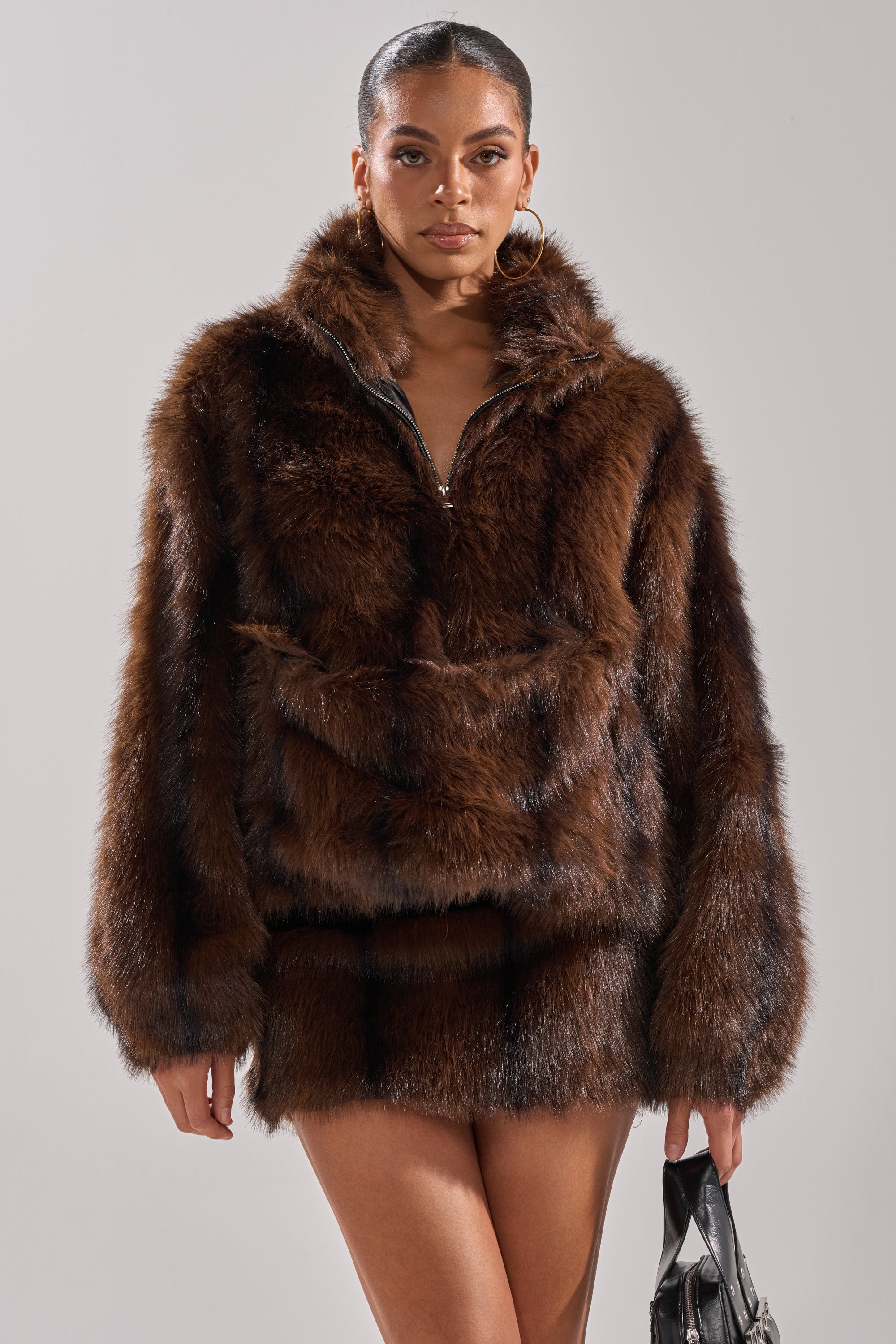 A woman models the COZY VIBES QUARTER ZIP FUR SWEATSHIRT in brown with a matching mini skirt, large hoop earrings, and a black handbag, standing against a plain light background.