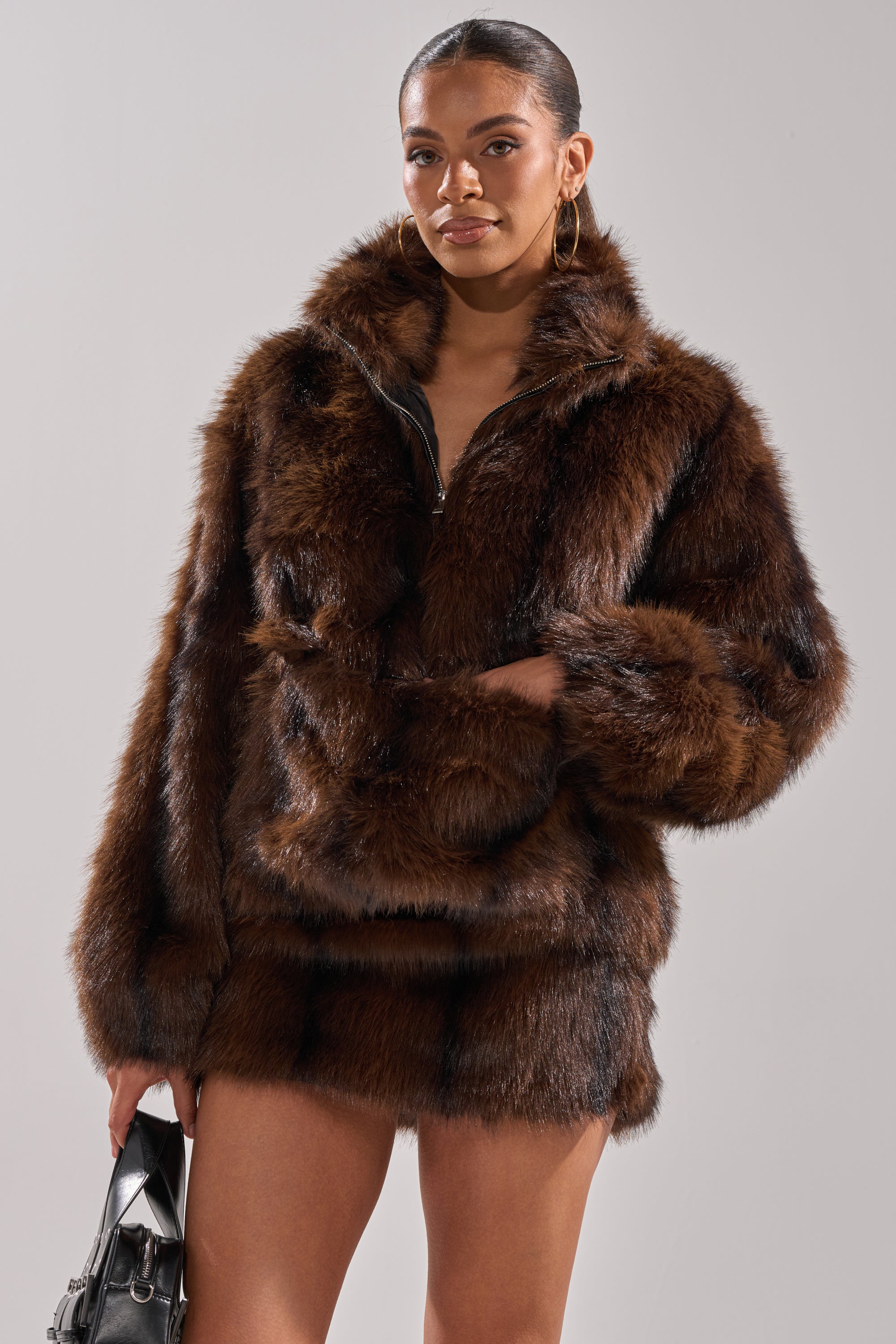 A woman models the COZY VIBES QUARTER ZIP FUR SWEATSHIRT and matching mini skirt, holding a small silver handbag against a plain background, exuding confidence.
