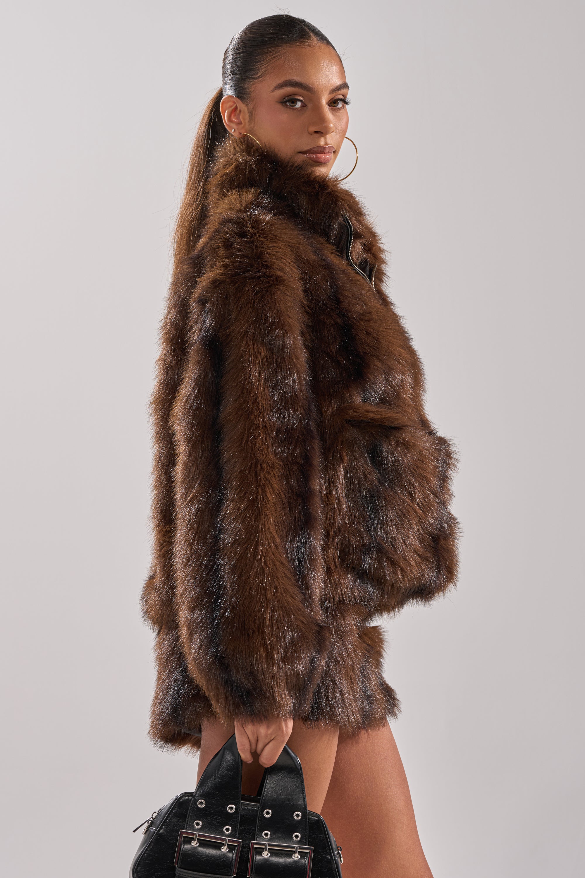 A woman with long, dark hair in a ponytail wears the COZY VIBES QUARTER ZIP FUR SWEATSHIRT and large hoop earrings, holding a black handbag against a plain light background.
