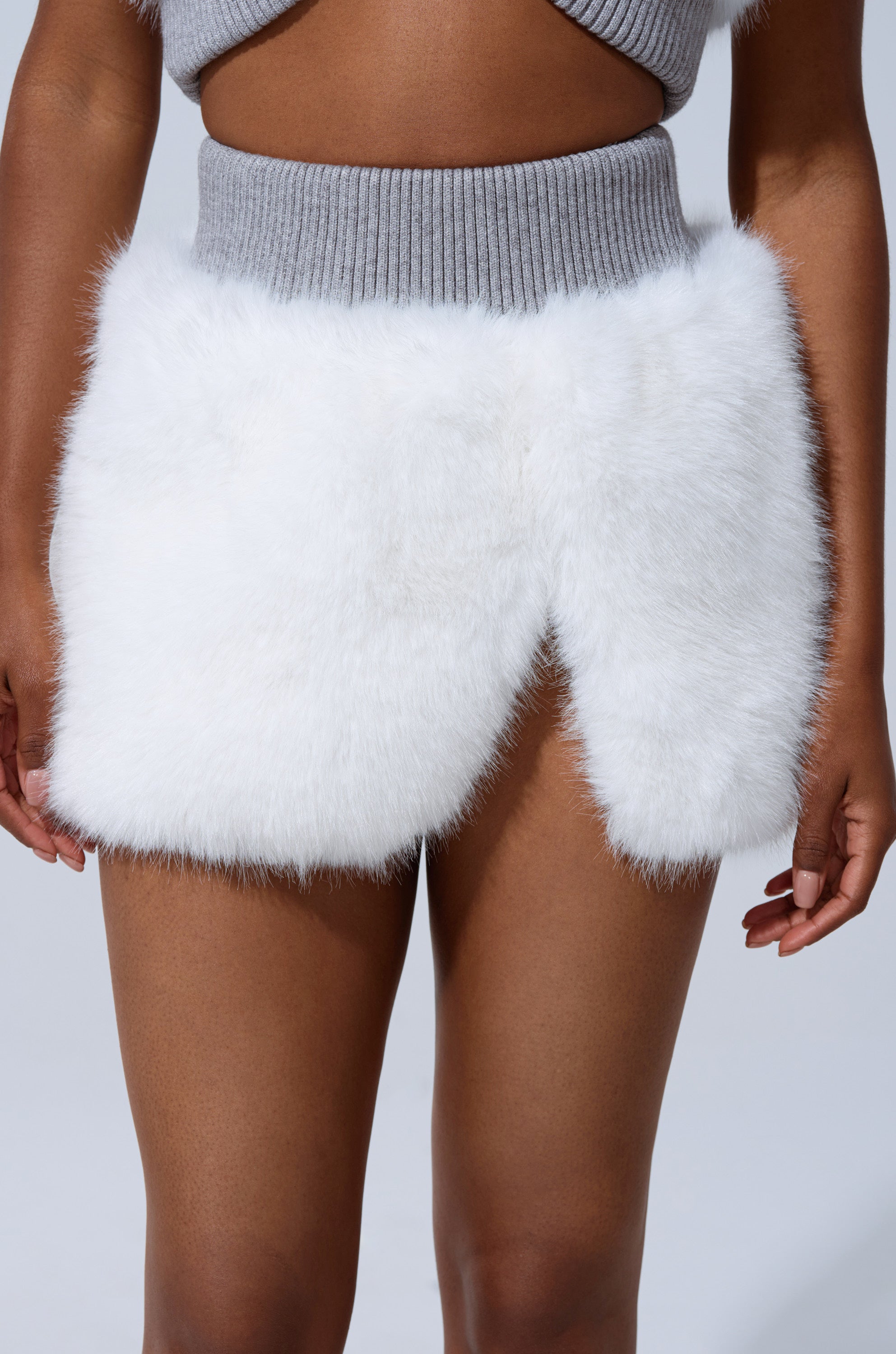 A person wearing a gray ribbed knit top and the COZY VIBES FUR MINI SKIRT—a white high-rise mini with faux fur texture and a front slit—photographed from waist to mid-thigh.