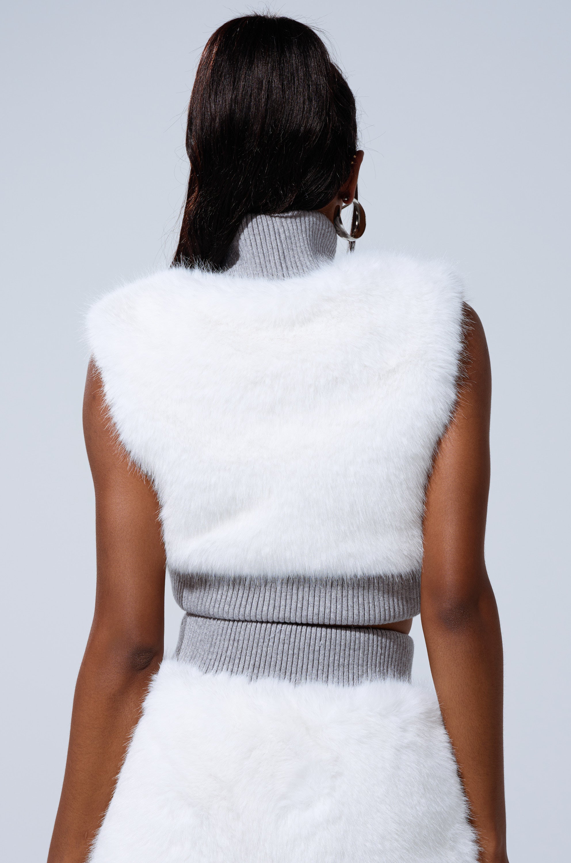 A person with long dark hair is seen from behind wearing the COZY VIBES FUR CARDIGAN, a sleeveless white faux fur piece with a high grey ribbed collar and matching waistband, creating a chic winter glam look against a light grey background.