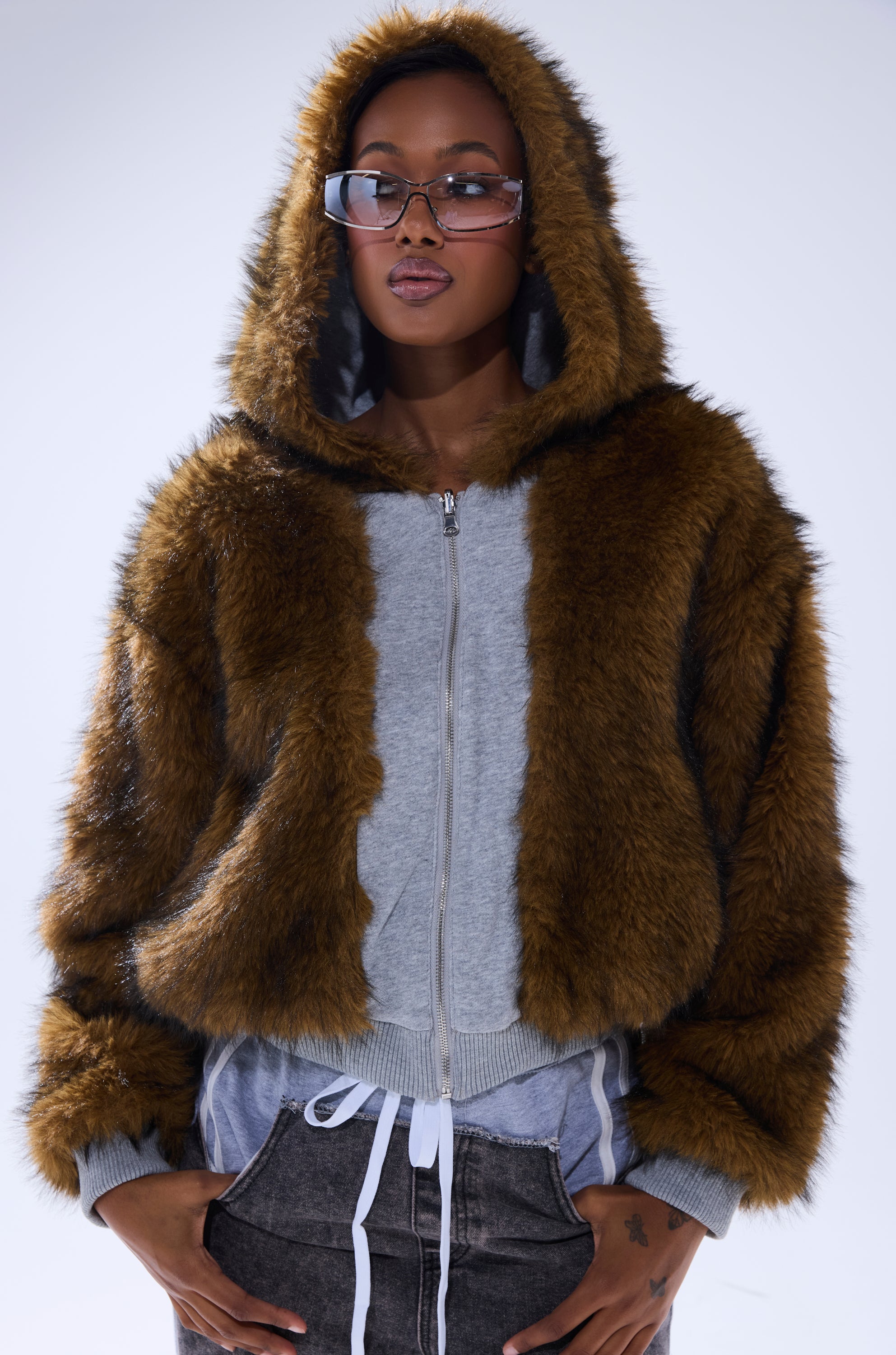 A person wears the BEST OF BOTH WORLDS REVERSIBLE FUR SWEATSHIRT—a versatile brown faux fur hooded jacket—over a gray zip-up hoodie, clear glasses, and dark jeans, standing against a plain light background.