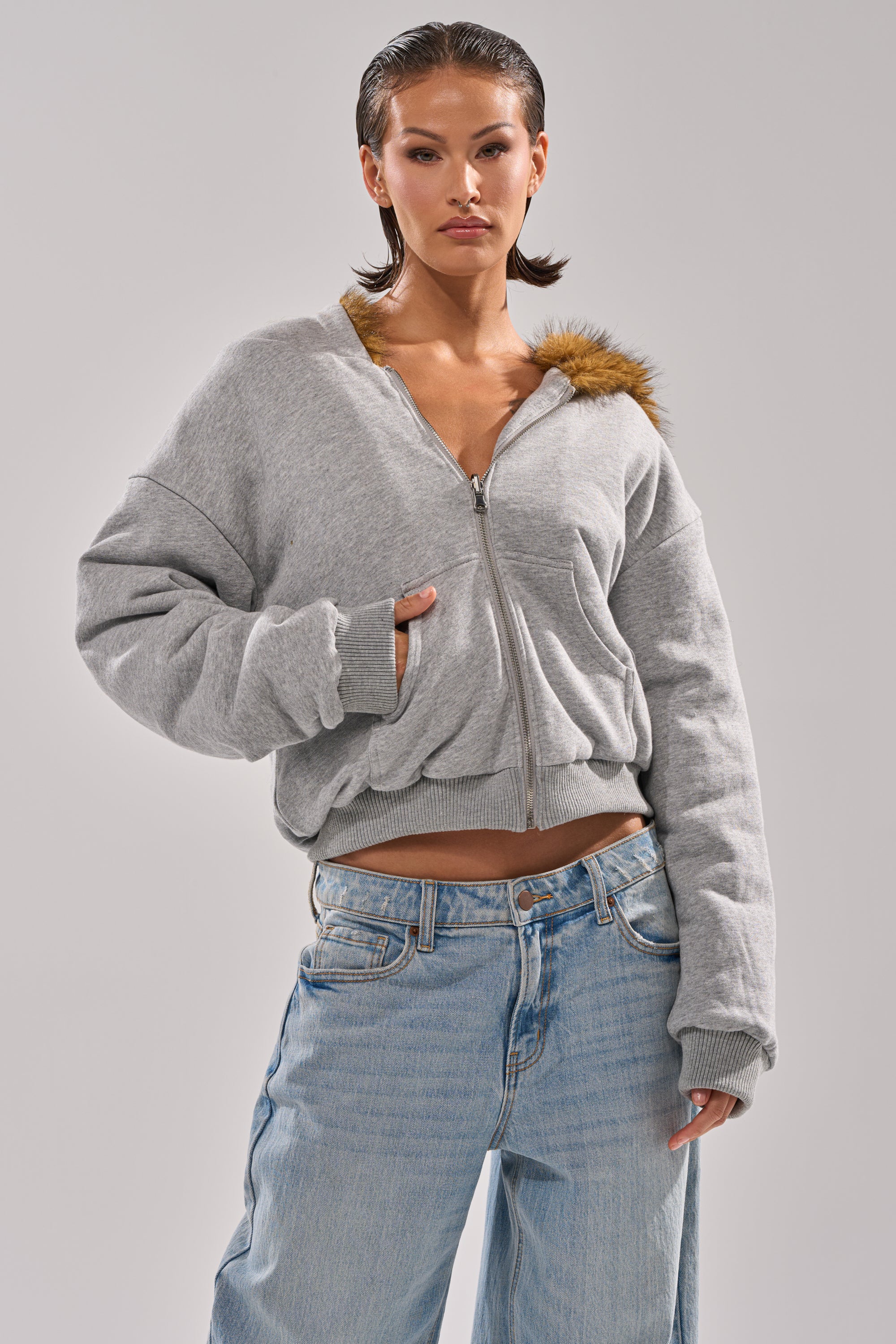 A person models the BEST OF BOTH WORLDS REVERSIBLE FUR SWEATSHIRT in gray with a zip-up front and light blue jeans, standing with one hand in their pocket against a plain background. 1
