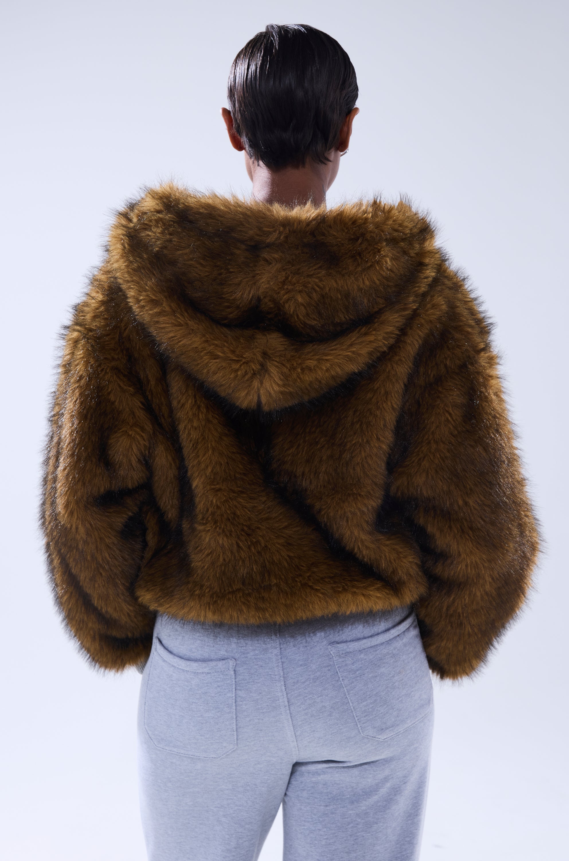 A person with short dark hair, seen from behind, wears the BEST OF BOTH WORLDS REVERSIBLE FUR SWEATSHIRT with a hood and light gray sweatpants against a plain light background.