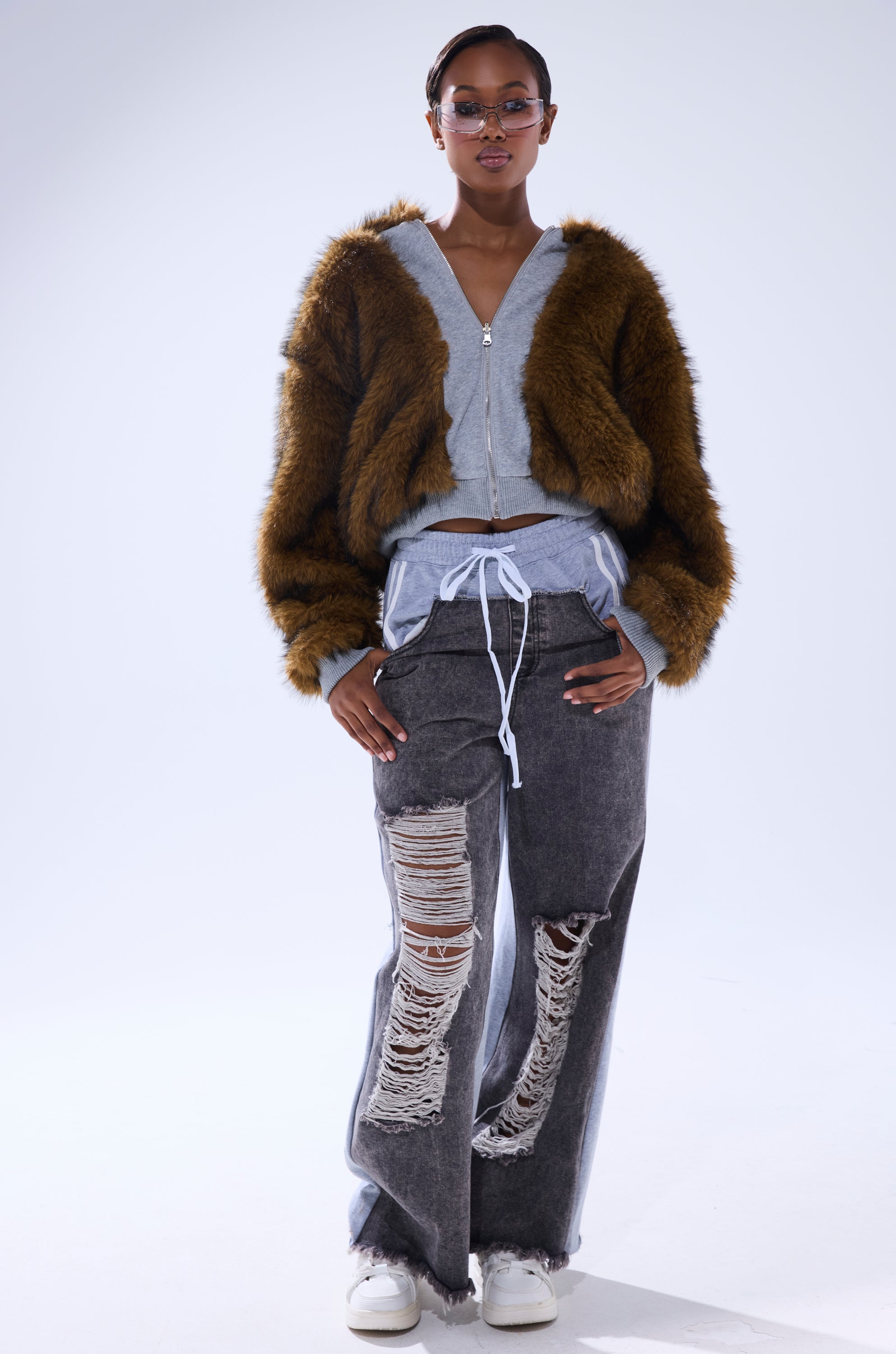A person in round sunglasses, the BEST OF BOTH WORLDS REVERSIBLE FUR SWEATSHIRT over a gray hoodie, distressed black jeans with large rips, and white sneakers stands confidently against a plain white background.