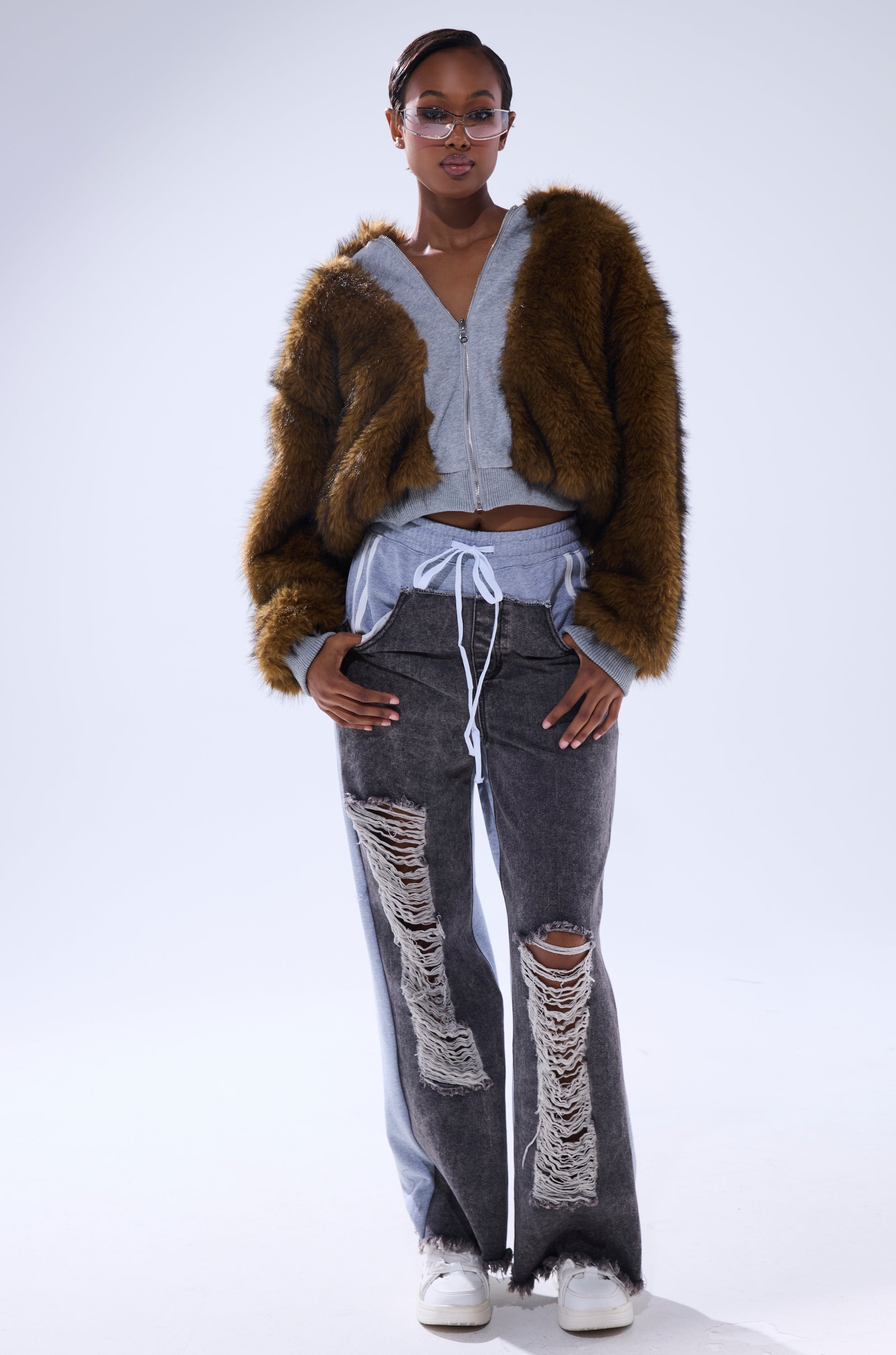 A person stands confidently wearing the BEST OF BOTH WORLDS REVERSIBLE FUR SWEATSHIRT in brown, paired with a light gray zip-up hoodie, distressed gray jeans with large rips, white sneakers, and glasses.
