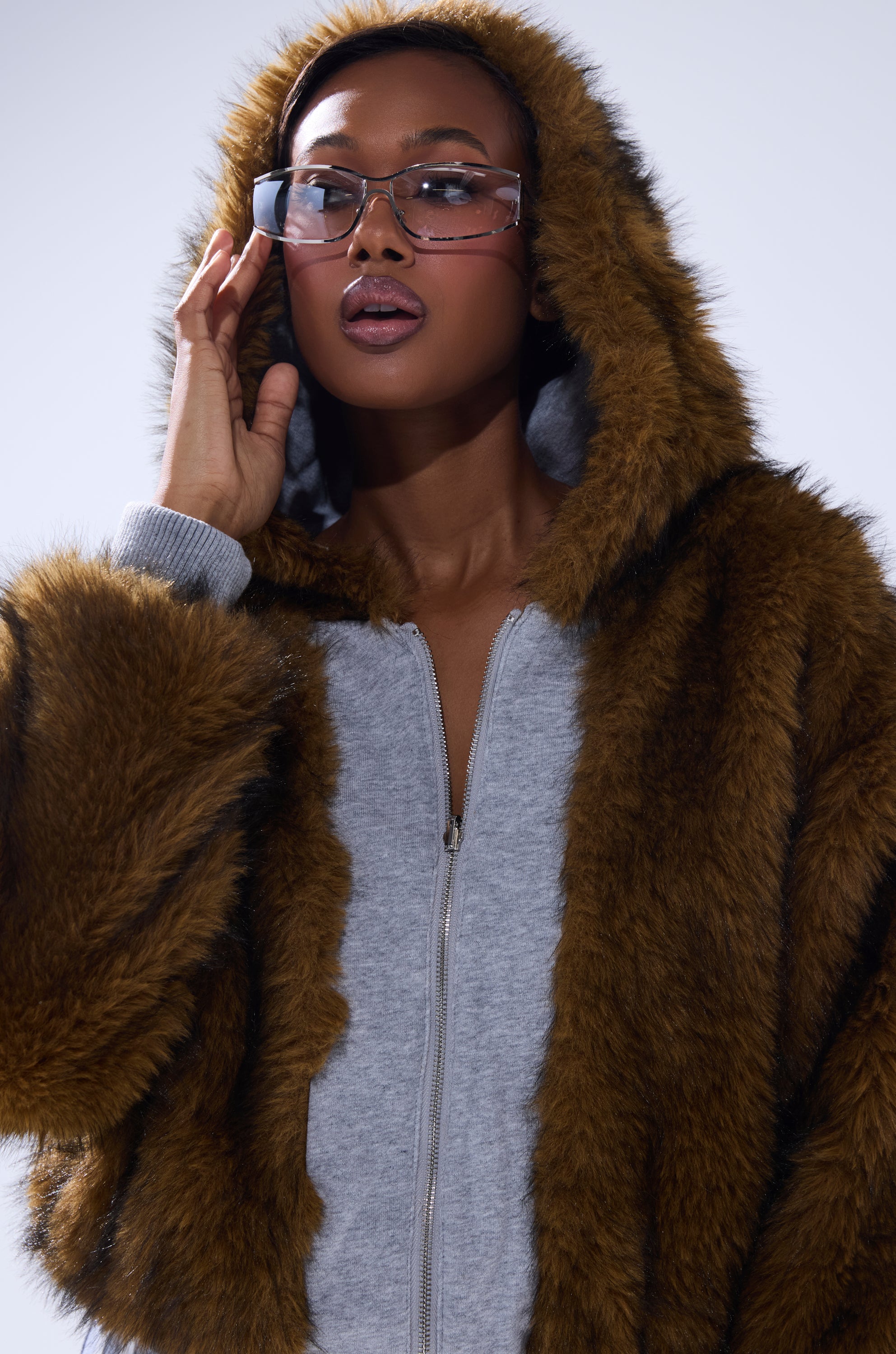 A person wears the BEST OF BOTH WORLDS REVERSIBLE FUR SWEATSHIRT in brown over a gray zip-up hoodie, touches clear-rimmed glasses, and looks slightly upward against a light background.