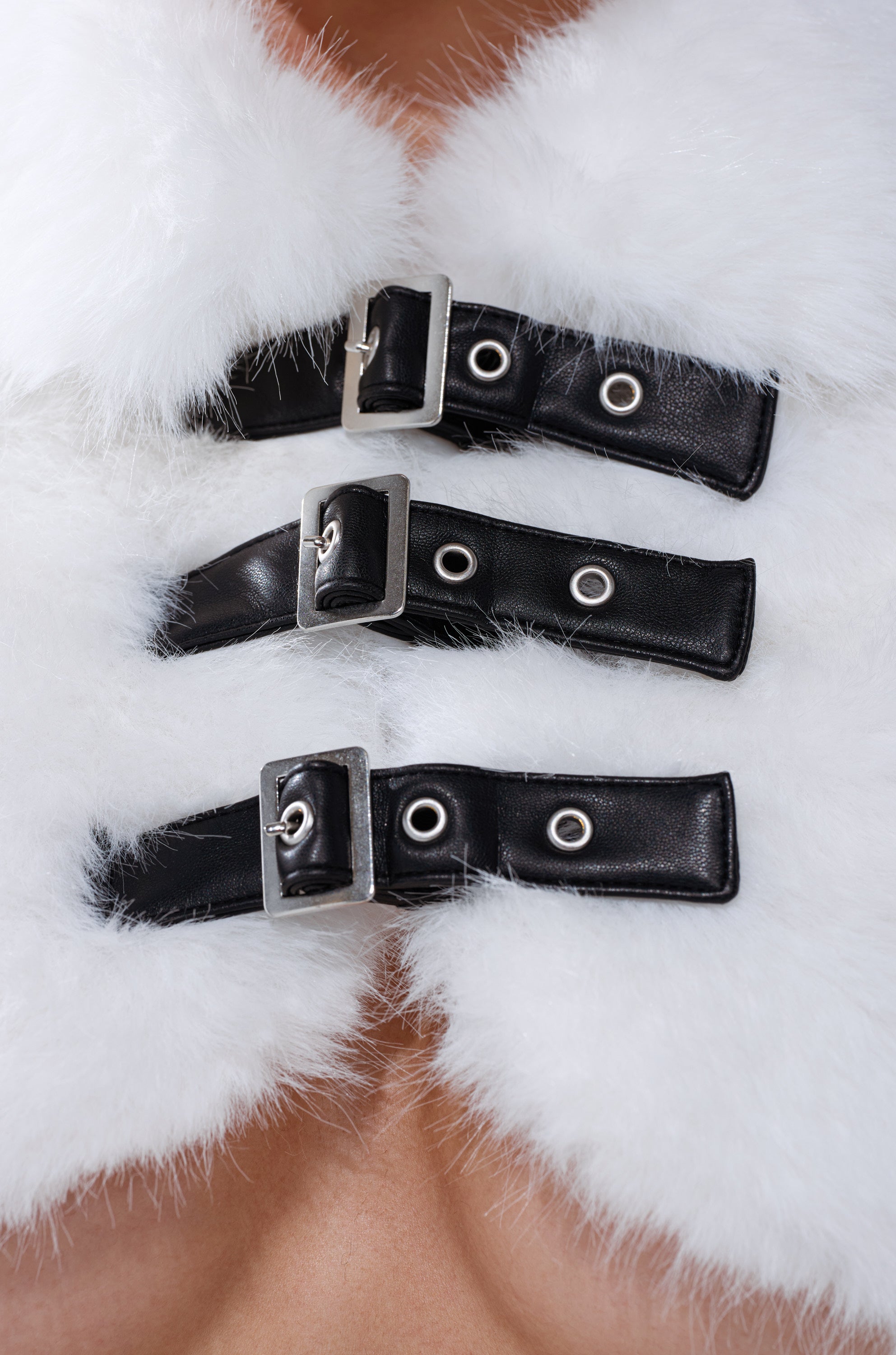 Close-up of three black leather straps with silver buckles fastened across the SNOW WHITE CROP FUR SWEATER's soft, fluffy faux fur, with a hint of bare skin in the background. 6
