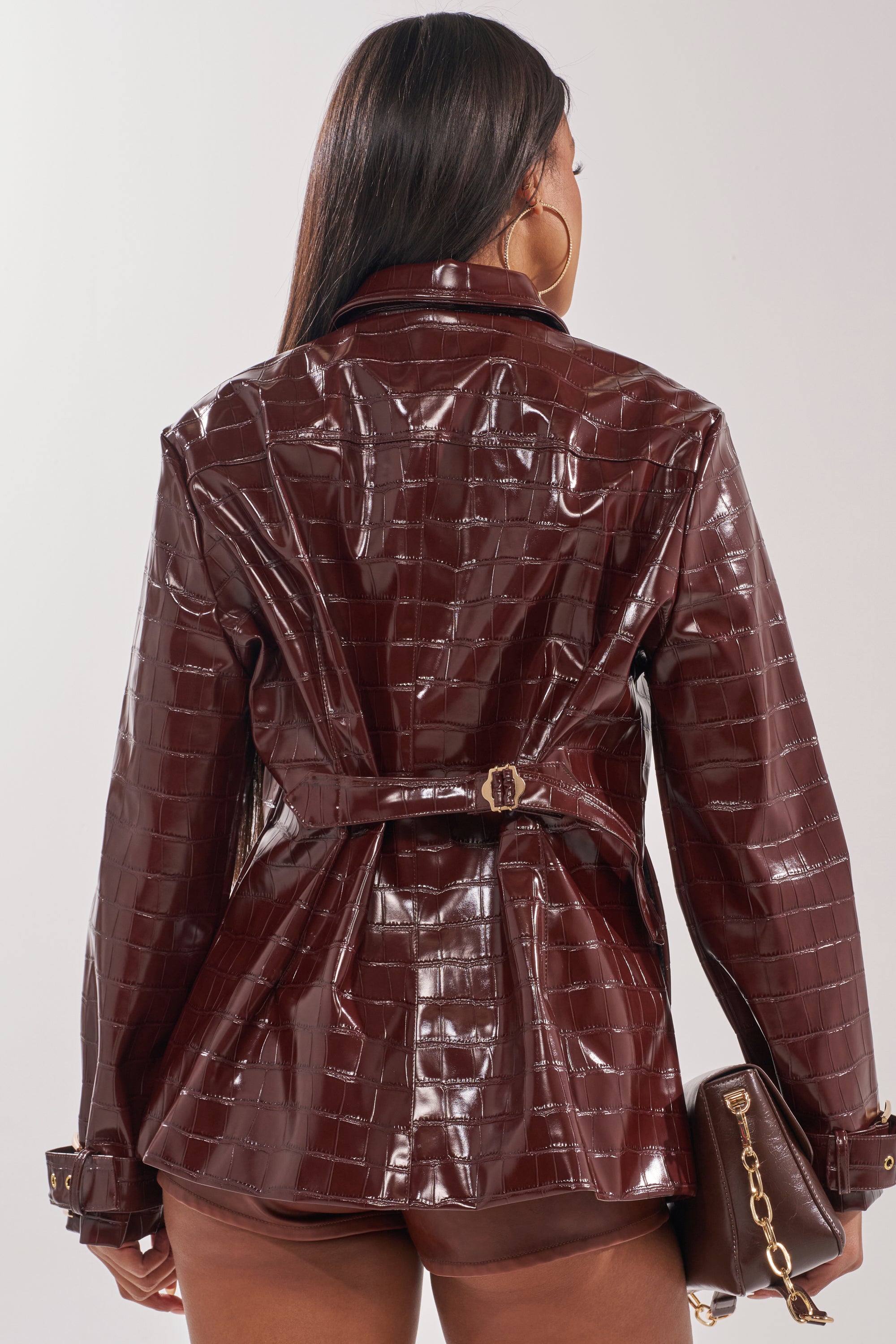 A woman with long straight hair, seen from behind, wears the glossy dark brown DONT STOP NOW FAUX LEATHER BUTTON DOWN jacket and holds a matching brown handbag with a gold chain. 1