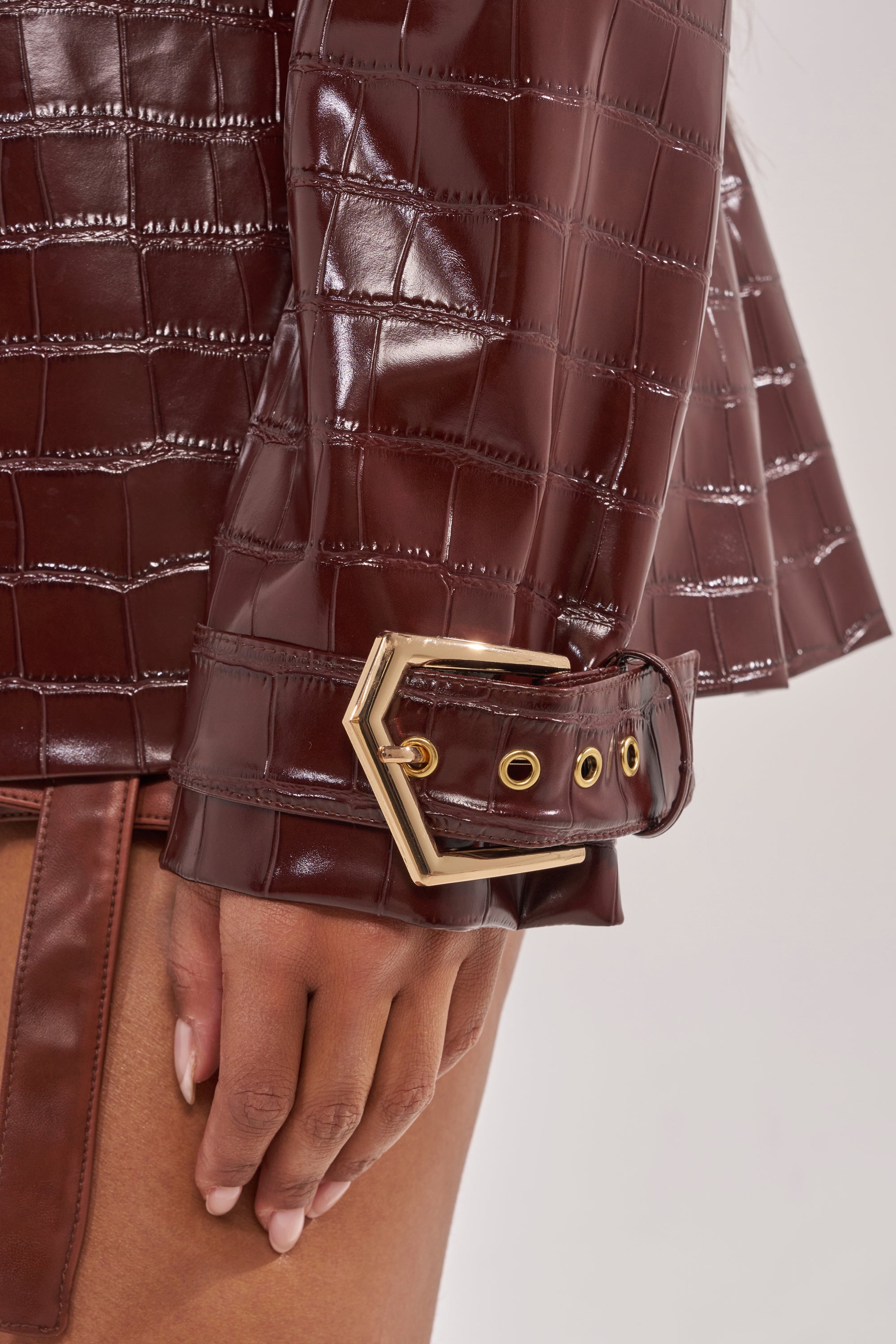 A close-up of a hand wearing the DONT STOP NOW FAUX LEATHER BUTTON DOWN, featuring shiny burgundy croc-embossed faux leather and a large gold hexagonal buckle at the sleeve cuff—a stylish pairing with a crisp shirt. 5