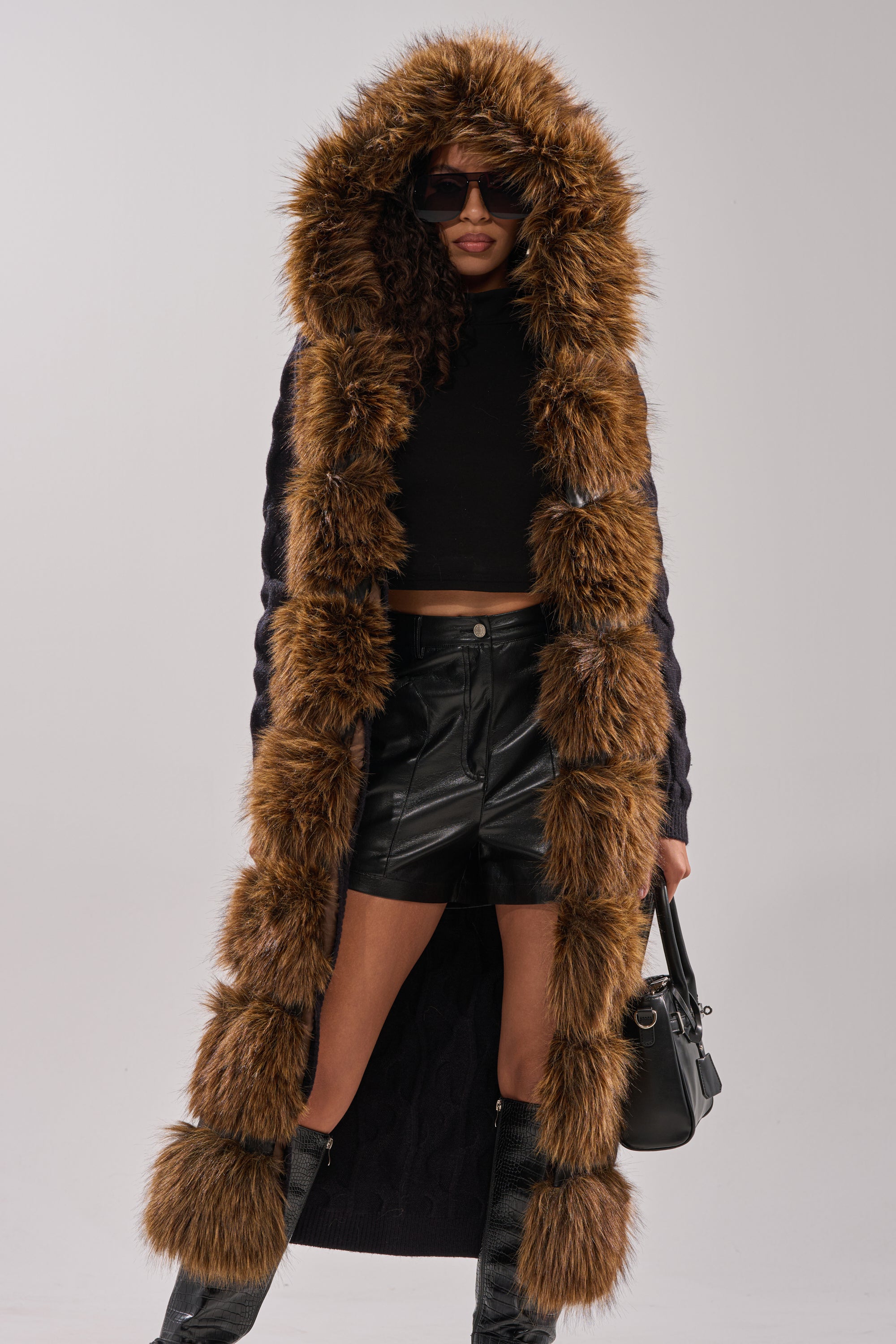 A woman exudes glamour in the WARM ME UP LONG CARDIGAN WITH FUR, styled with a black crop top, leather mini skirt, knee-high boots, sunglasses, and a black handbag. 0