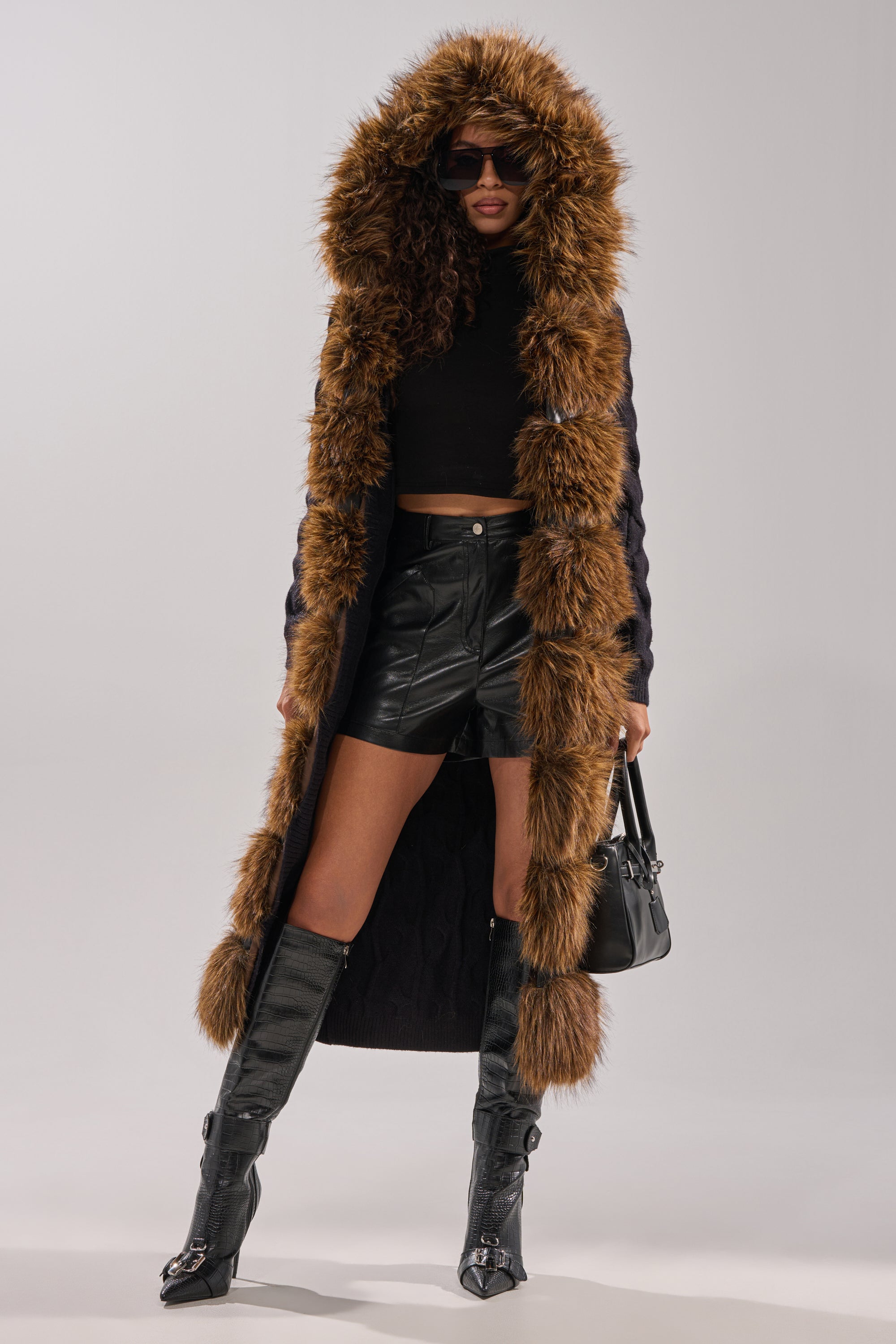 A woman stands confidently against a light grey background, wearing the WARM ME UP LONG CARDIGAN WITH FUR, paired with a black crop top, leather shorts, high boots, sunglasses, and holding a black handbag. 9