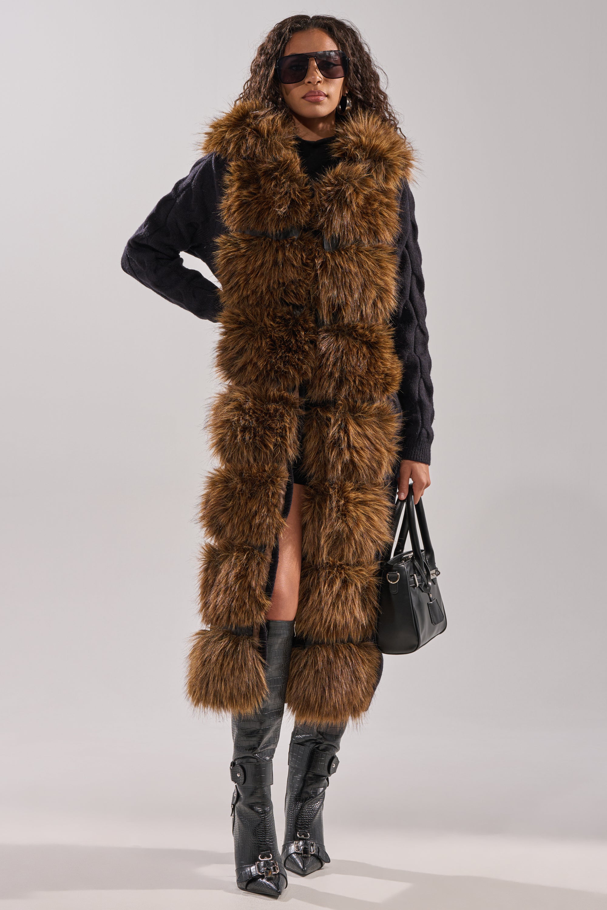 A woman stands confidently against a neutral background, wearing the WARM ME UP LONG CARDIGAN WITH FUR over a black outfit, accessorized with sunglasses, a black handbag, and knee-high boots with buckles. 10
