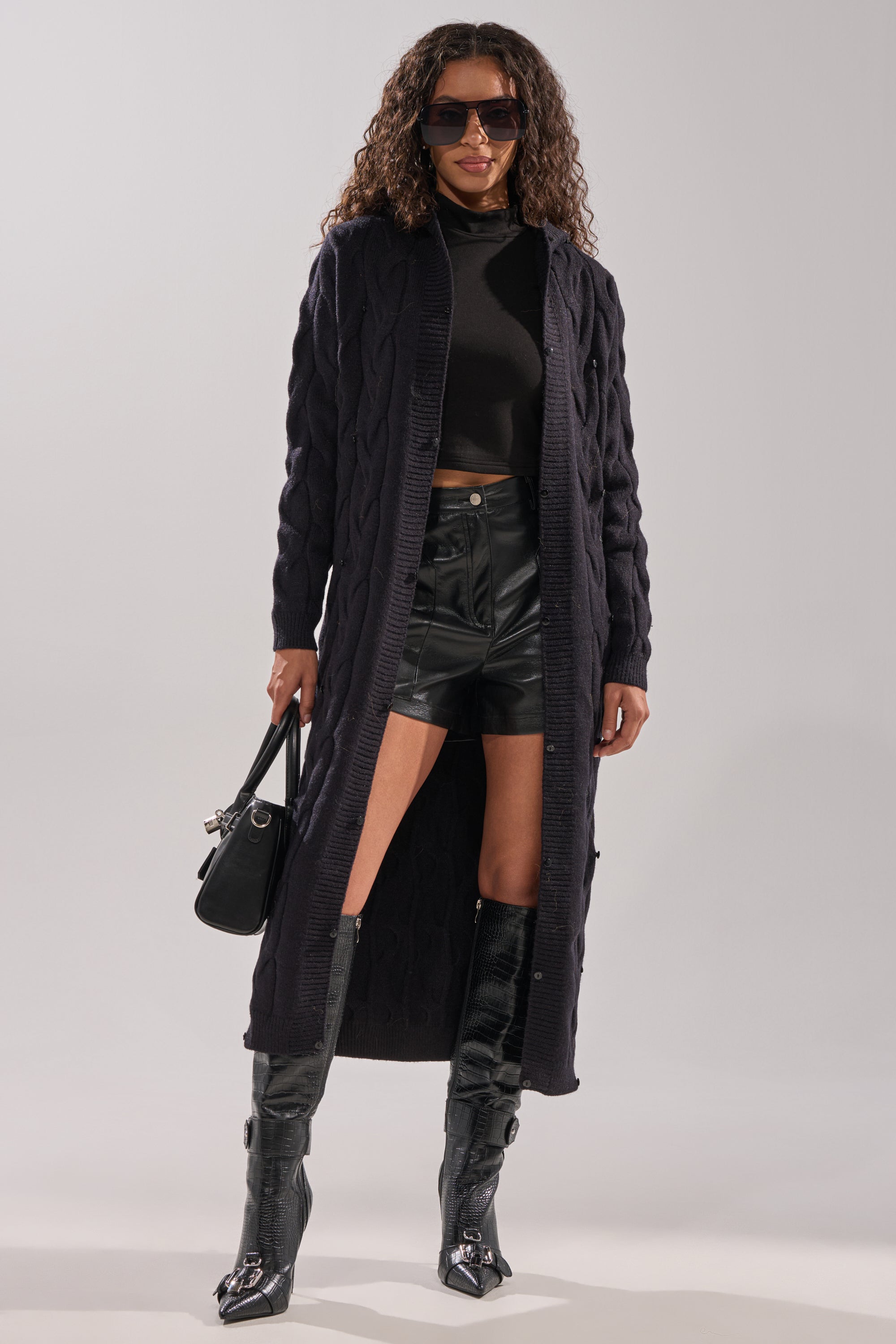 A woman stands confidently against a light background wearing sunglasses, knee-high boots, a black handbag, leather shorts, and the WARM ME UP LONG CARDIGAN WITH FUR over a black turtleneck crop top. 13