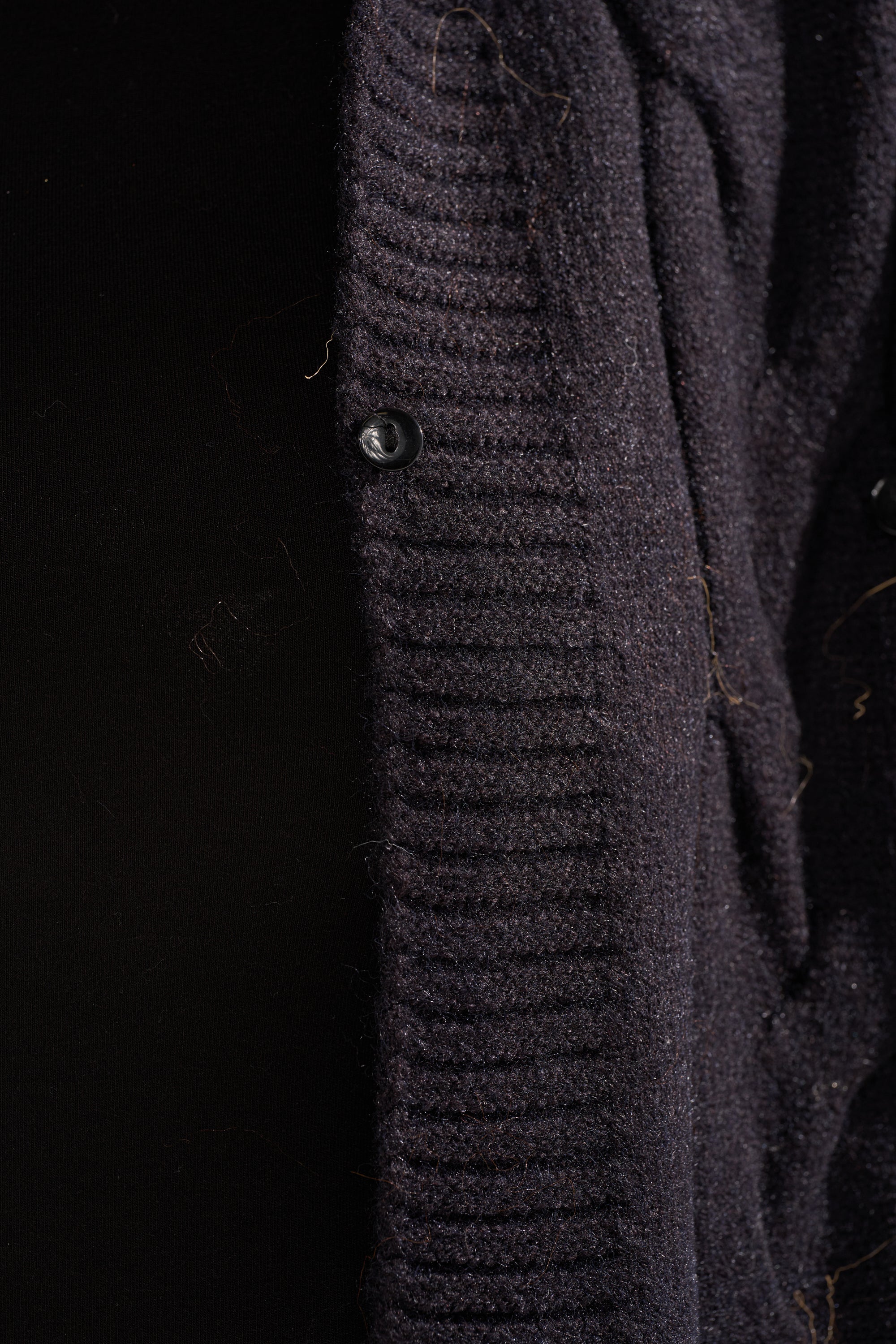 Close-up of the WARM ME UP LONG CARDIGAN WITH FUR, a dark knit design with fur details, shown partially unbuttoned against a black background. 15