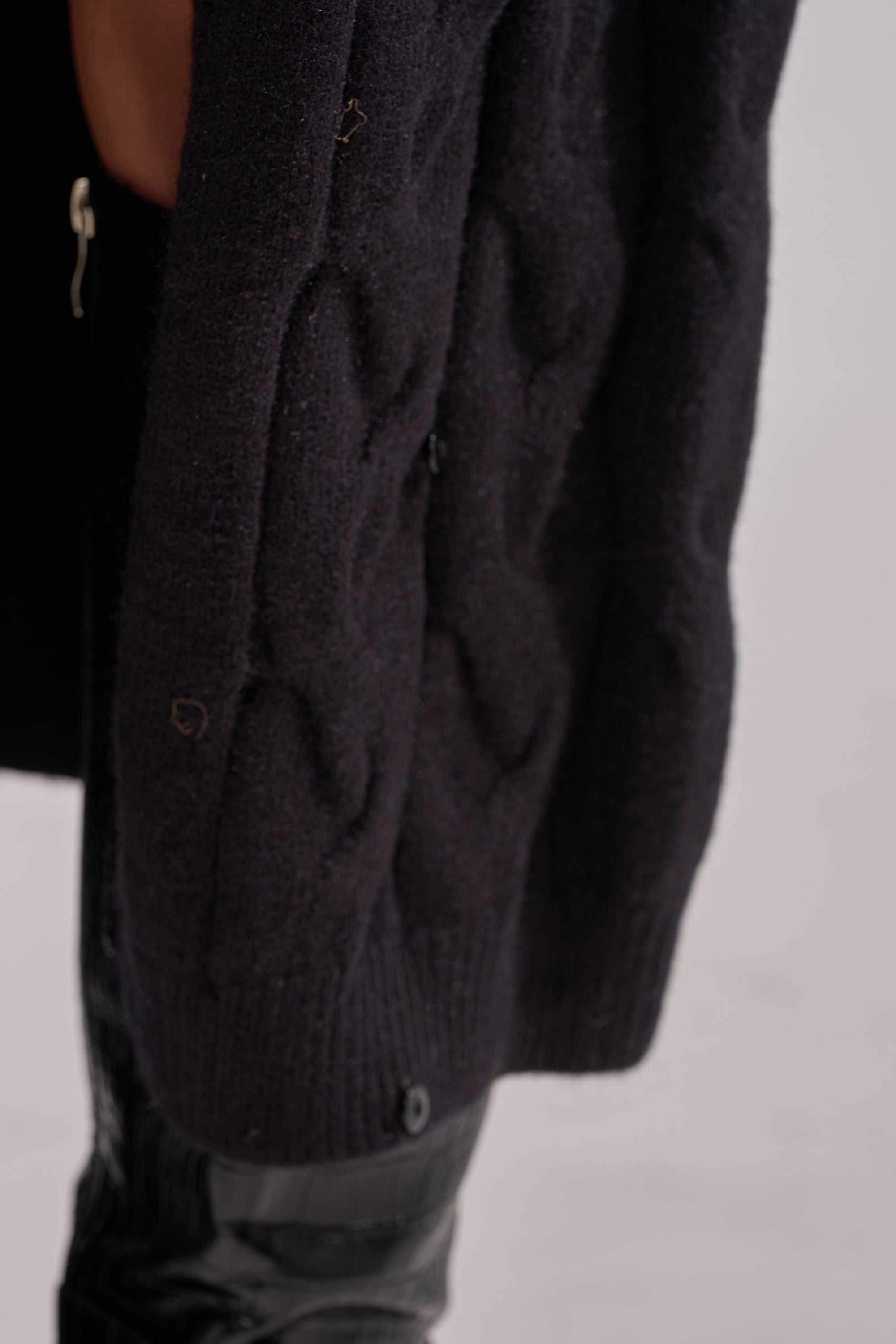 Close-up of the WARM ME UP LONG CARDIGAN WITH FUR, a dark cable knit layer with thick texture, draped over a person in black pants and boots. The neutral background is softly blurred. 16