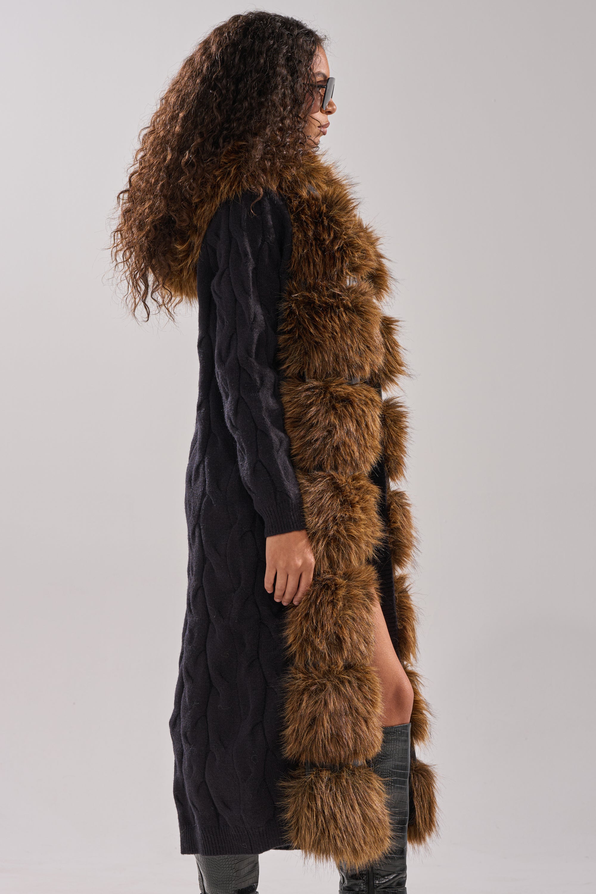 A woman with curly hair and sunglasses stands in profile against a light gray background, wearing the WARM ME UP LONG CARDIGAN WITH FUR— a black cable knit design featuring fur trim and a hood—paired with a brown faux fur stole. 5