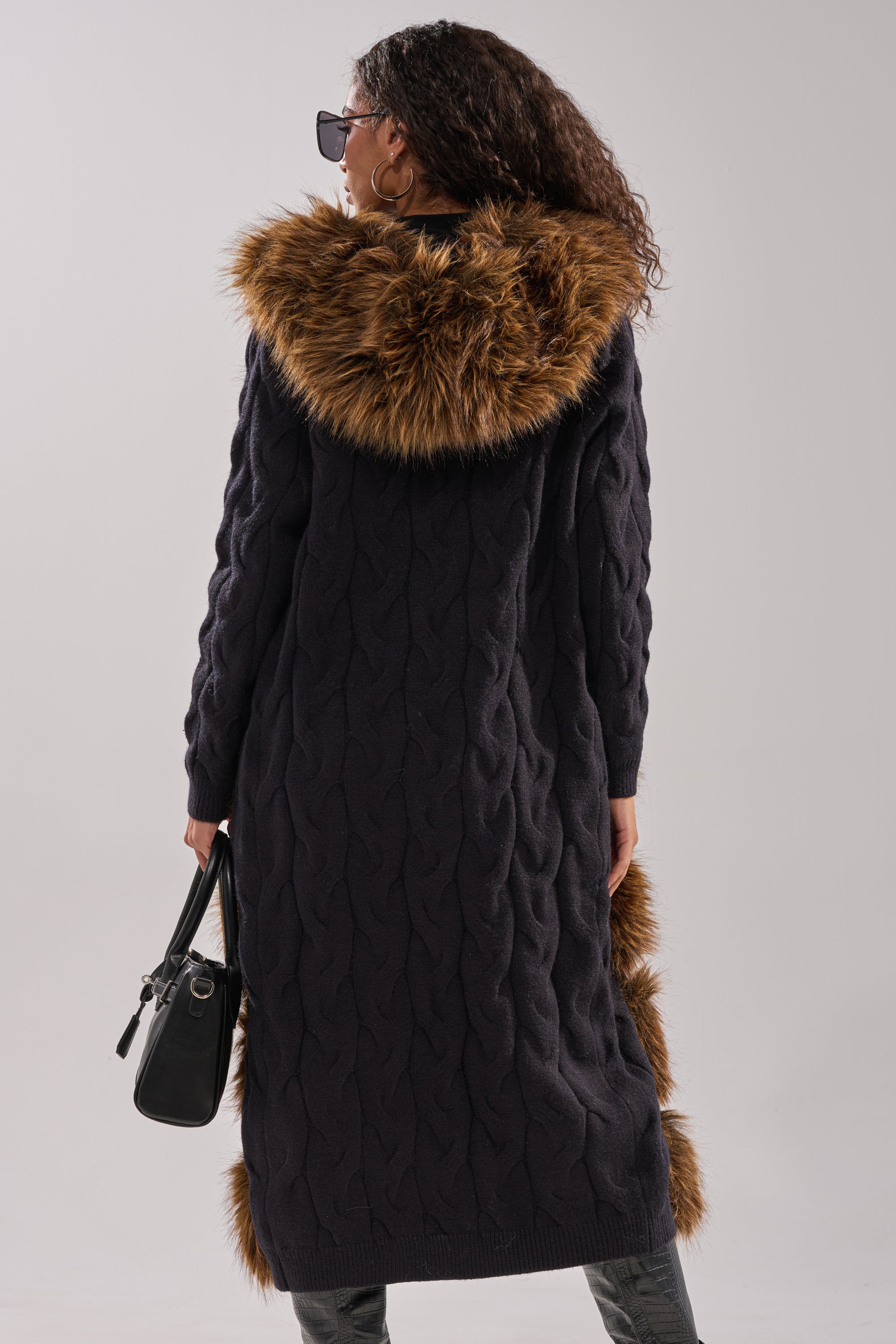 A woman with curly hair wears sunglasses and the WARM ME UP LONG CARDIGAN WITH FUR, featuring a brown faux fur hood. She holds a black handbag and is photographed from the back against a plain background. 6
