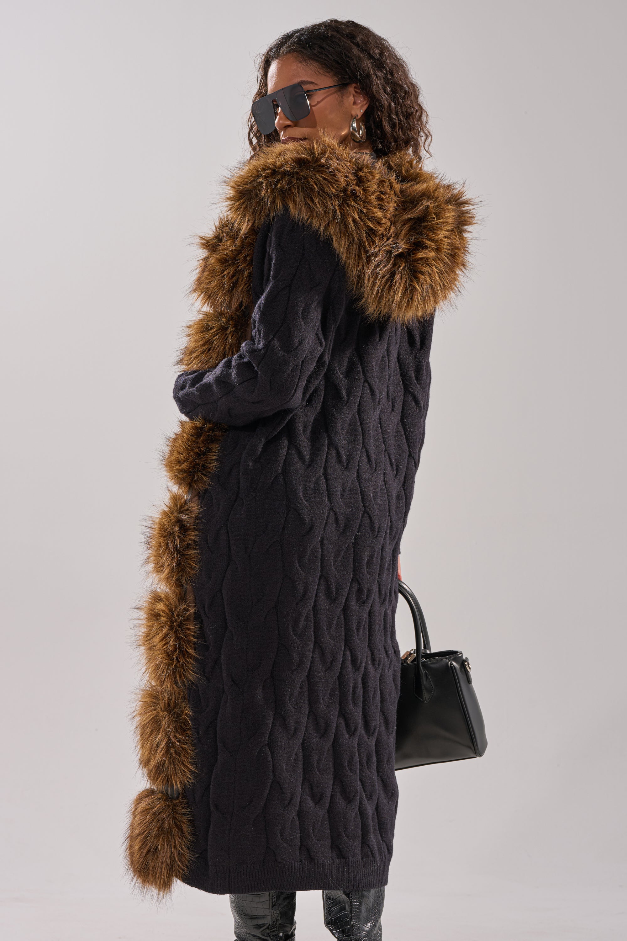 A person wears the WARM ME UP LONG CARDIGAN WITH FUR, styled with a black handbag and large sunglasses, standing against a plain background with their back to the camera. 7