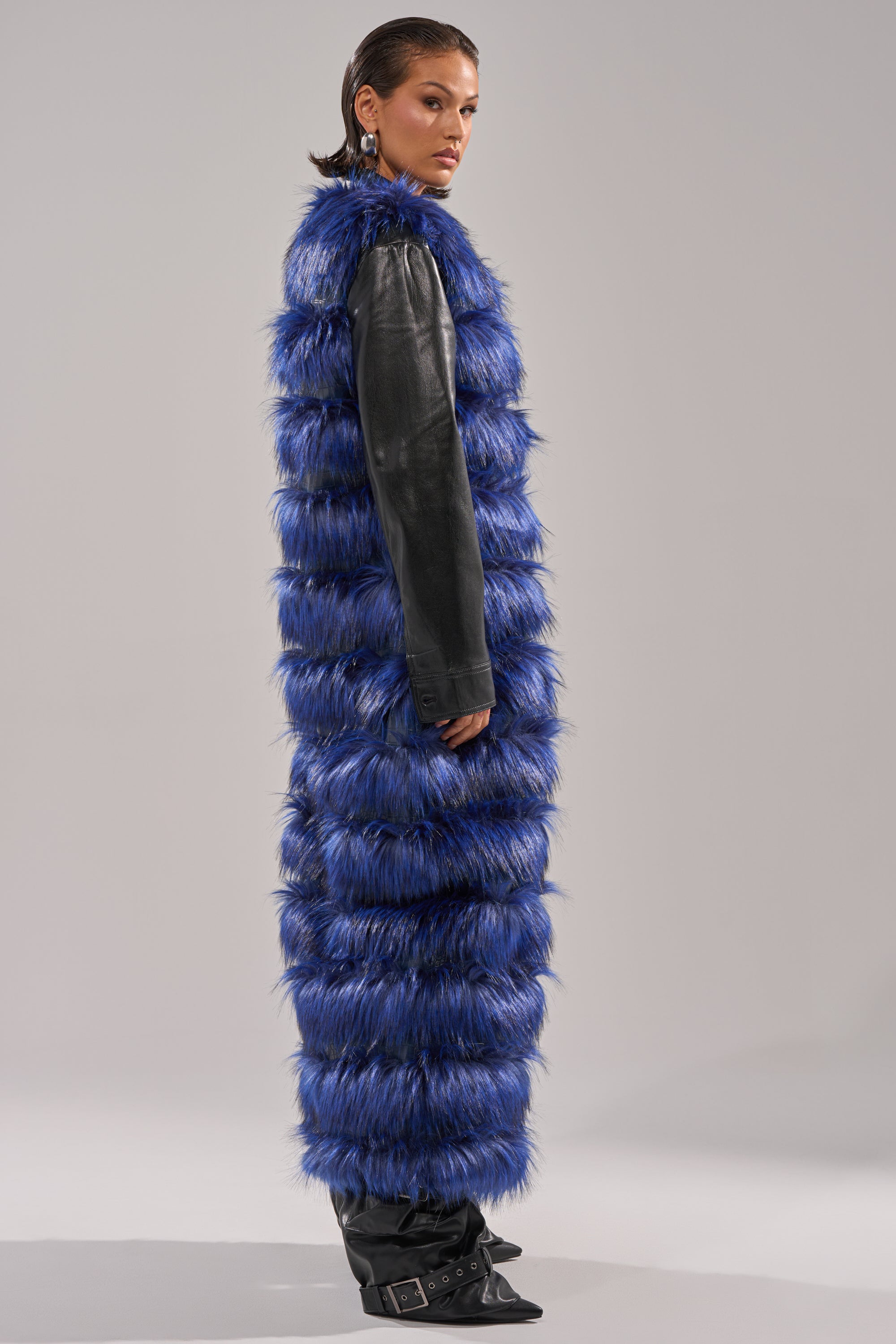 A person stands sideways wearing the RIZZA PANELED FUR VEST in blue over a black leather top and black boots, set against a plain light gray background.