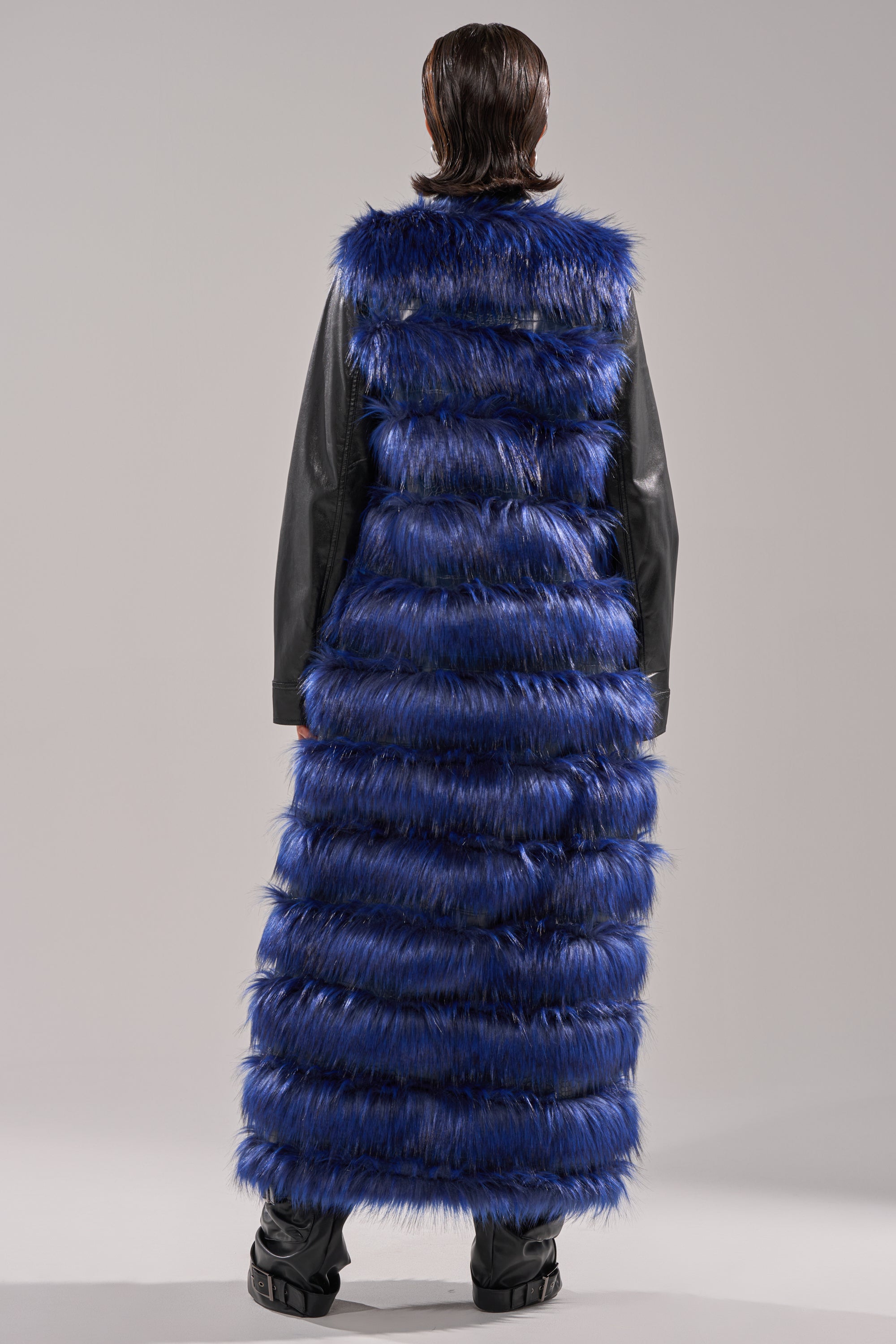 A person stands with their back turned, wearing the RIZZA PANELED FUR VEST in blue—a longline, layered faux fur vest reaching the ankles—over a black outfit against a plain light gray background.