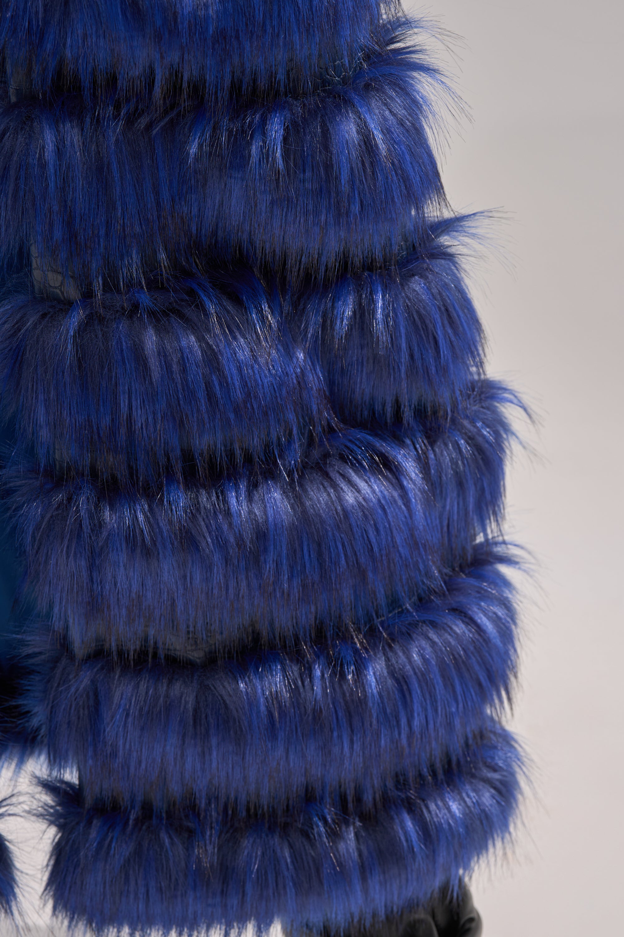 Close-up of the RIZZA PANELED FUR VEST, a blue longline vest crafted from bright faux fur with horizontal, layered sections, highlighting its soft and fluffy texture against a neutral background.