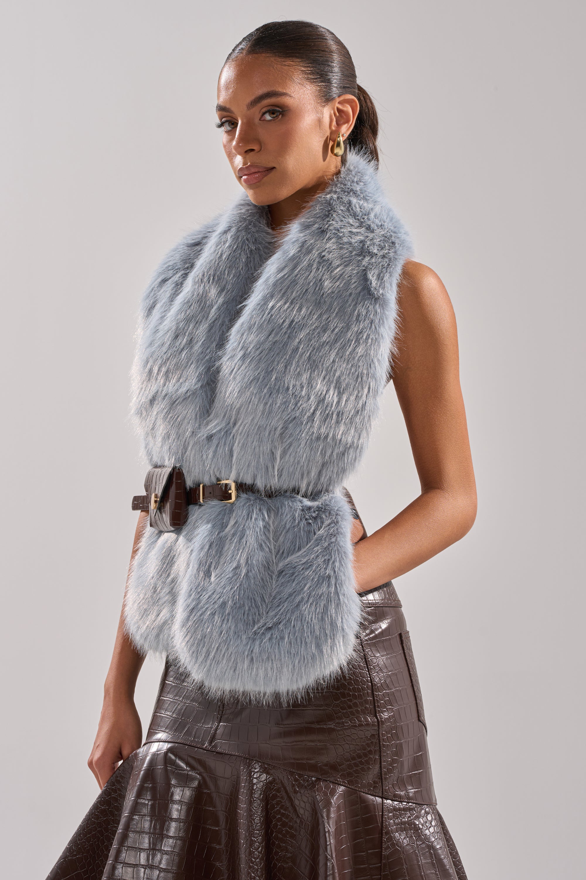 A woman wears the ITS GIVING FUR BLOUSE, a sleeveless light blue faux fur top with an adjustable belt, paired with a brown crocodile-textured skirt. She stands confidently against a plain background—a chic winter essential. 1